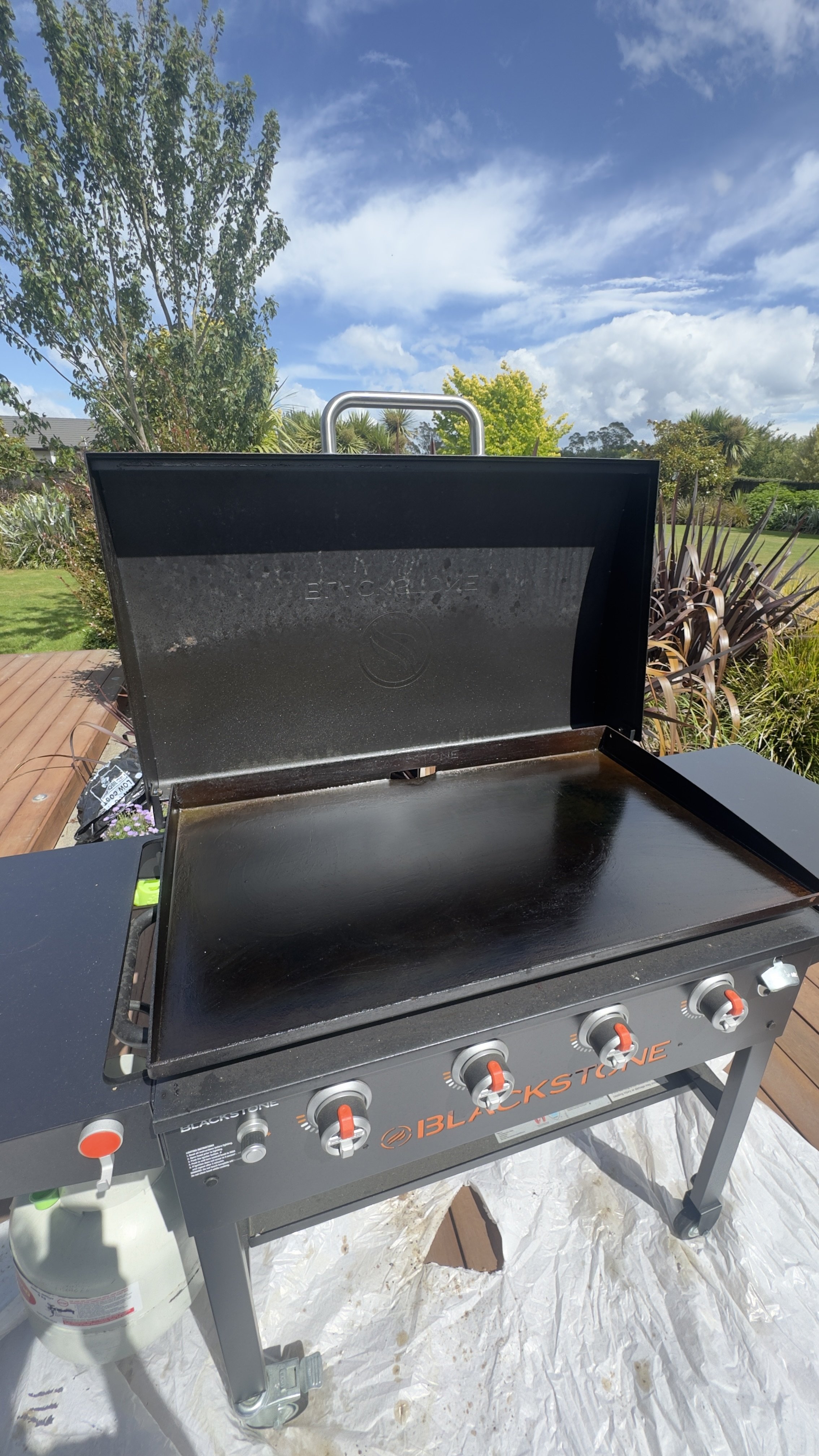 Blackstone gas grill with lid open, situated outdoors on a wooden deck, with trees, plants, and a blue sky with clouds in the background.