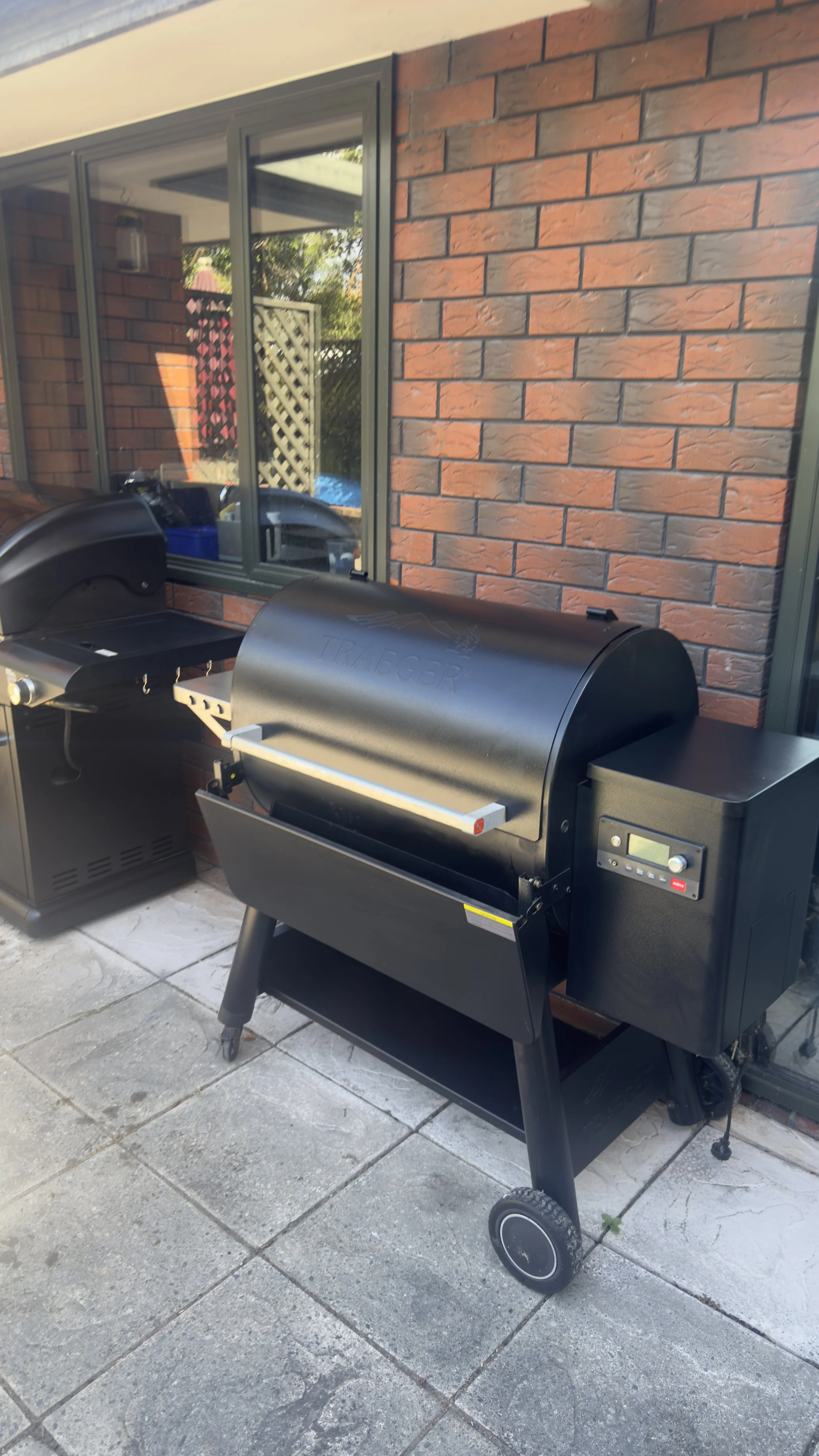 A black Traeger pellet grill on a patio beside a brick wall, with another grill to the left and a window reflecting a backyard with trees and fencing.