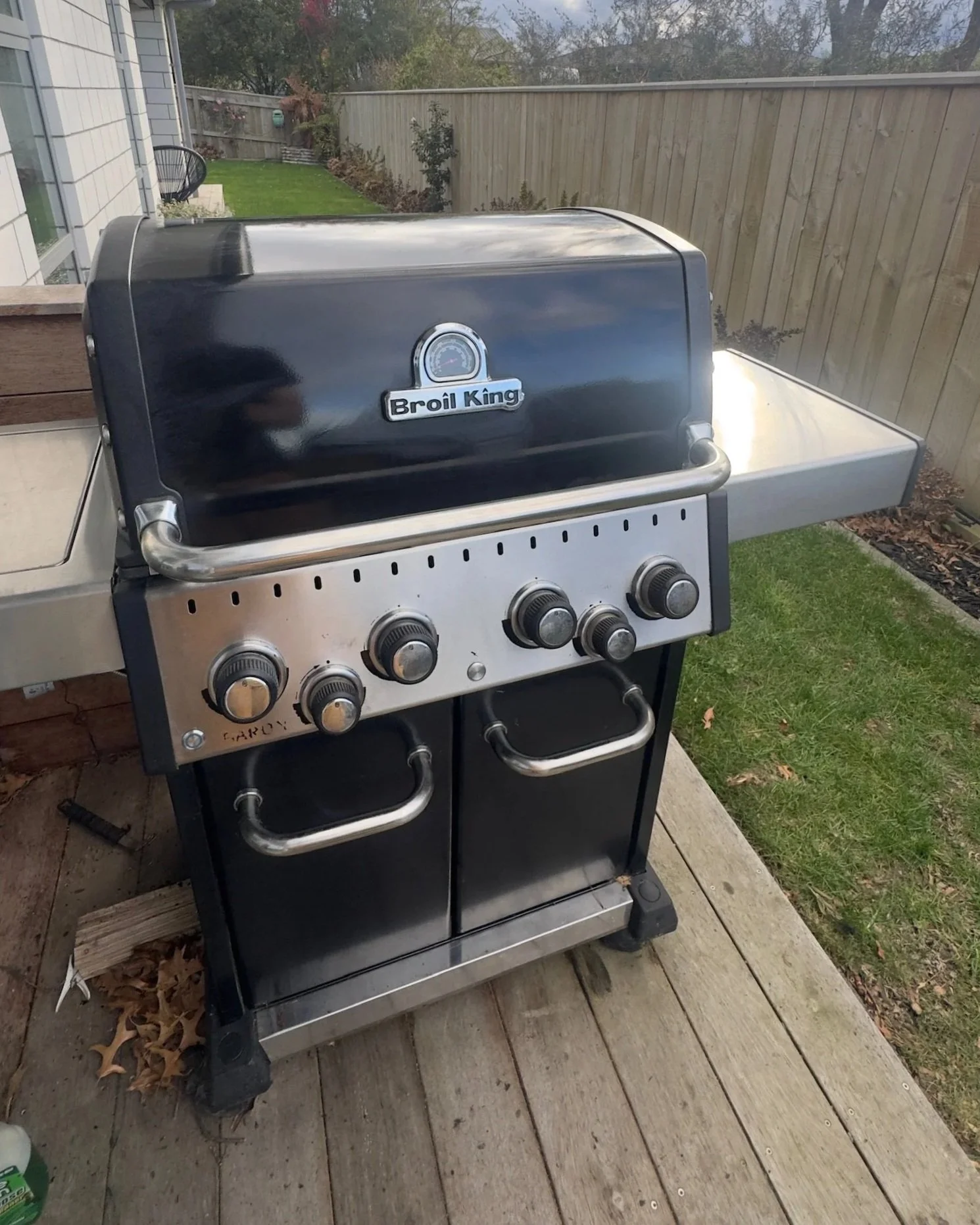 A black and silver Broil King gas grill on a wooden deck in a backyard, with a grassy lawn, a wooden fence, and trees in the background.