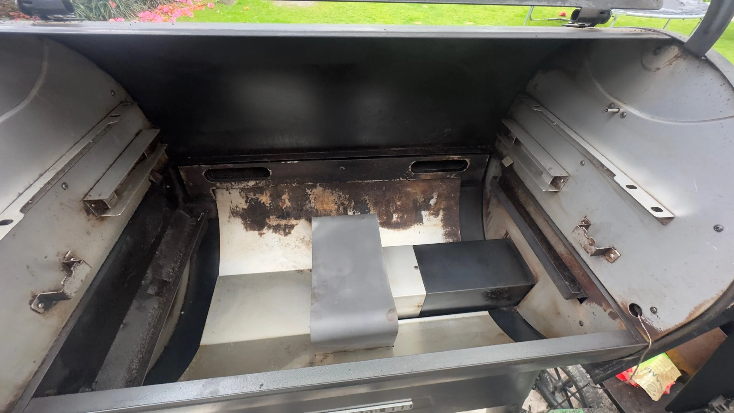 Interior view of an empty, rusty, and worn-out gas grill with some blackened areas and missing parts, outdoors with a lawn and flowers in the background.