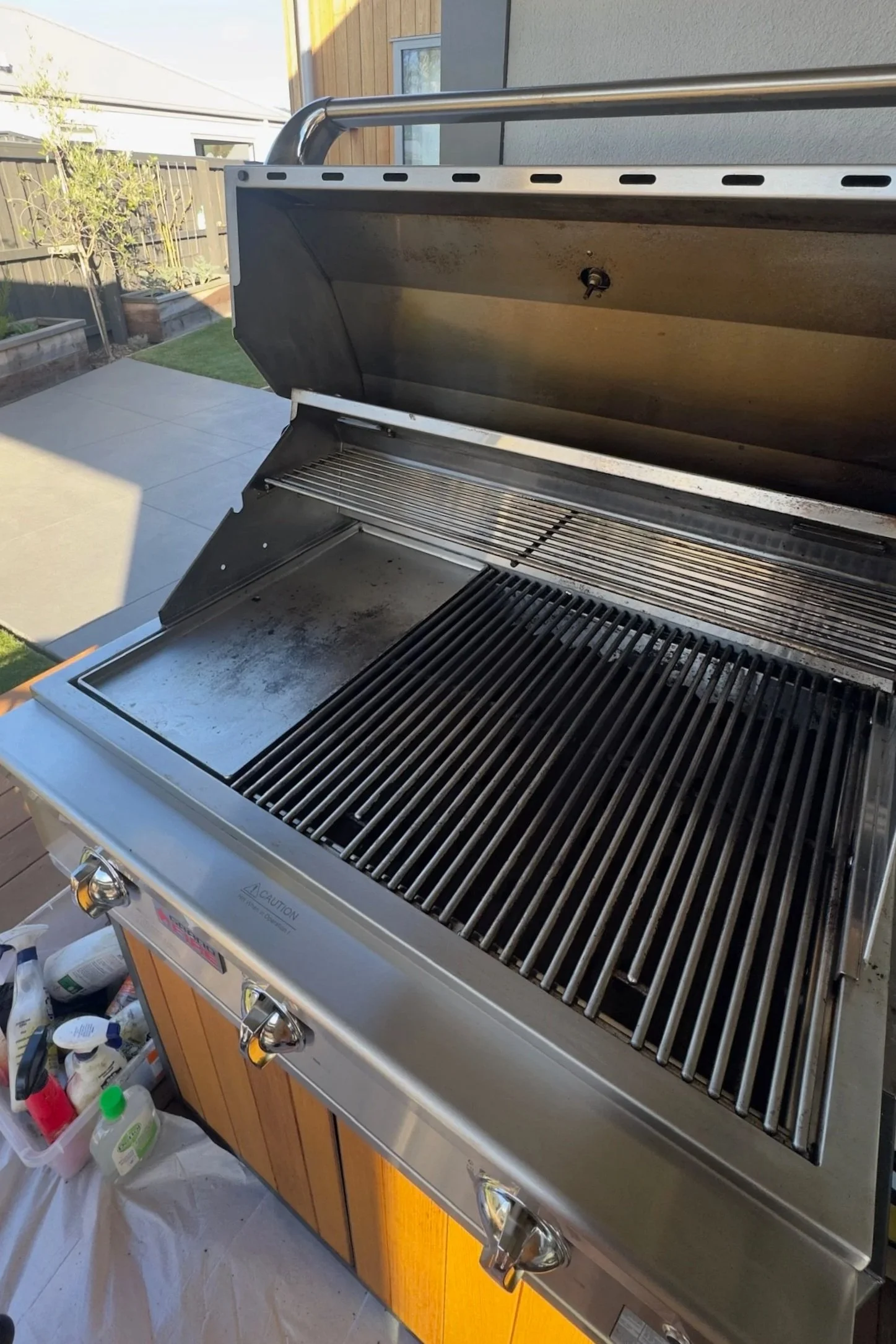 A stainless steel barbecue grill with a wooden cabinet underneath, located outdoors. The grill has a lid that is open, revealing the cooking grates inside. Various bottles and cleaning supplies are visible below the grill.