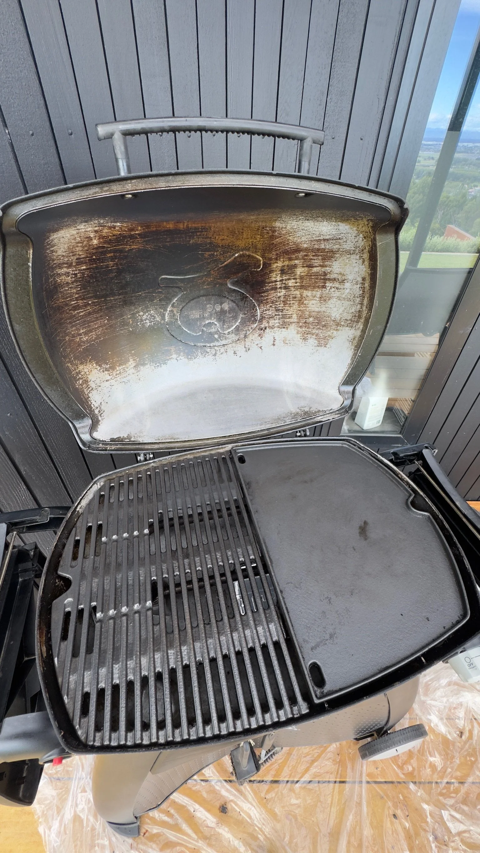 An open electric grill with a scratched and greasy cooking surface and a clean grilling grate in a modern outdoor setting.