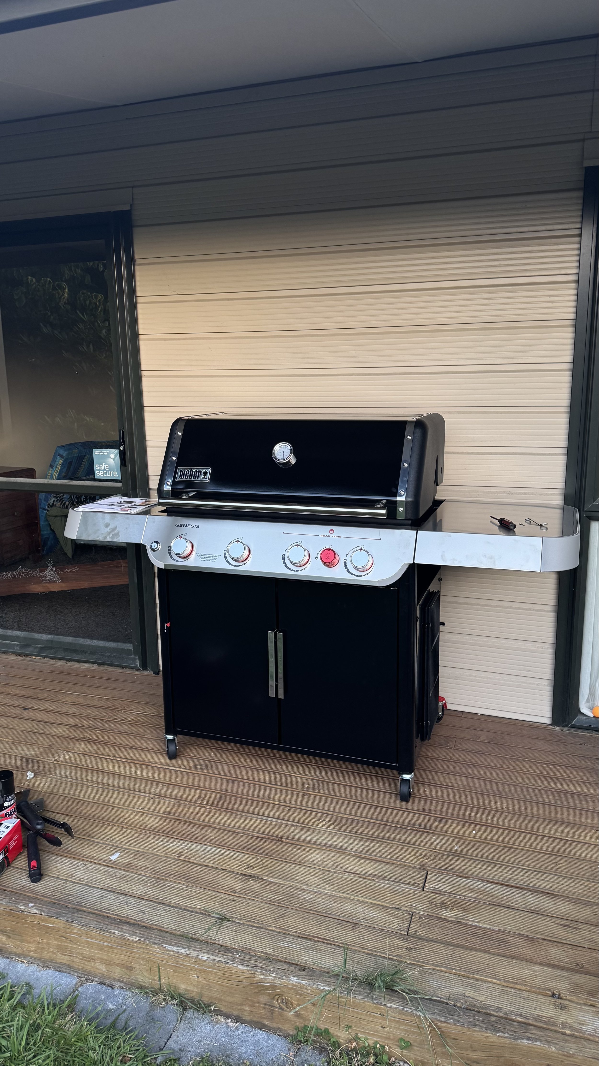 Black propane gas grill with a closed lid, four control knobs, and stored under a white folding table on a wooden deck outside a house.