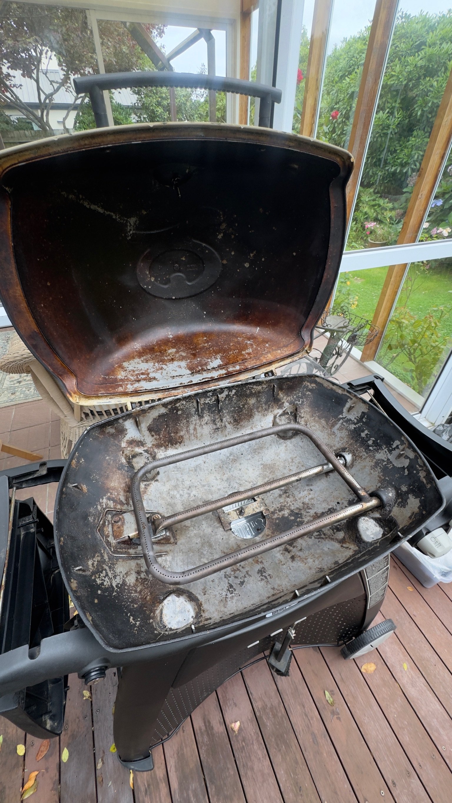 A barbecue grill with the lid open in a screened porch area, showing a dirty, rusted cooking surface, with some grease and grime. The grill is located on a wooden deck with fallen leaves visible on the floor. Outside, there are plants and flowers.