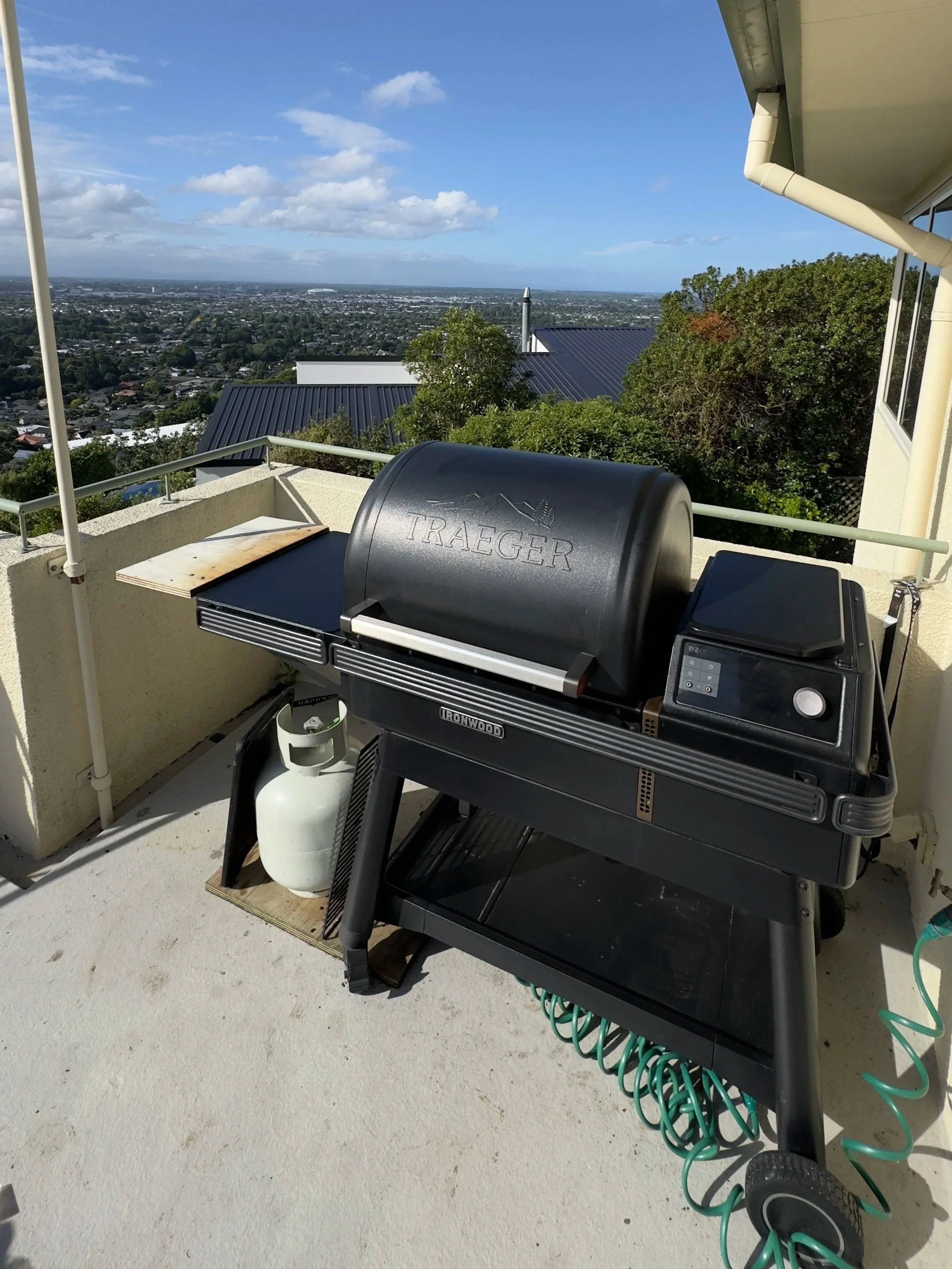 A black Traeger grill on a balcony with a propane tank underneath, overlooking a cityscape with houses, trees, and a blue sky with clouds.