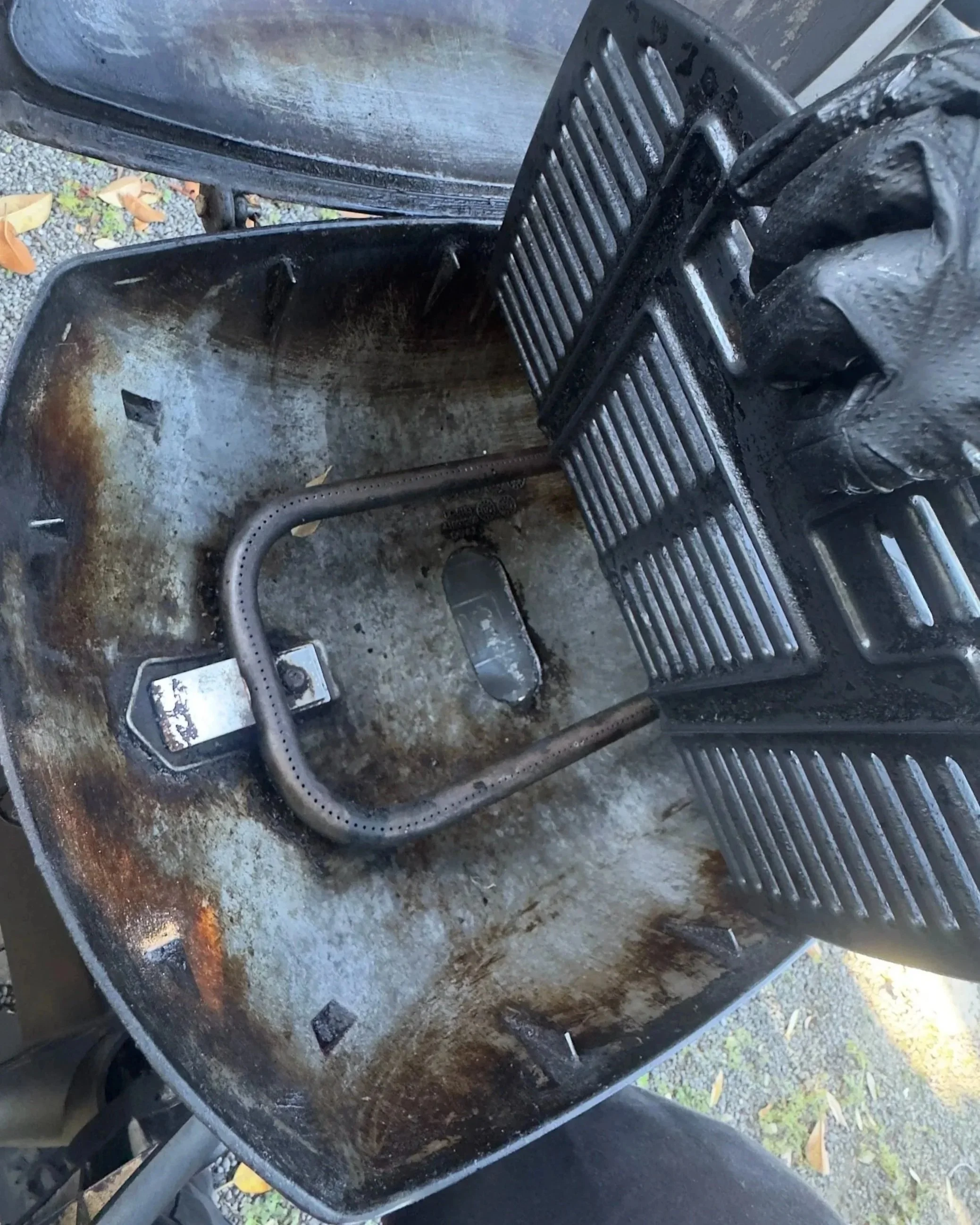 Inside of a rusty outdoor grill or smoker with a grate leaning against the side, no food or charcoal present.