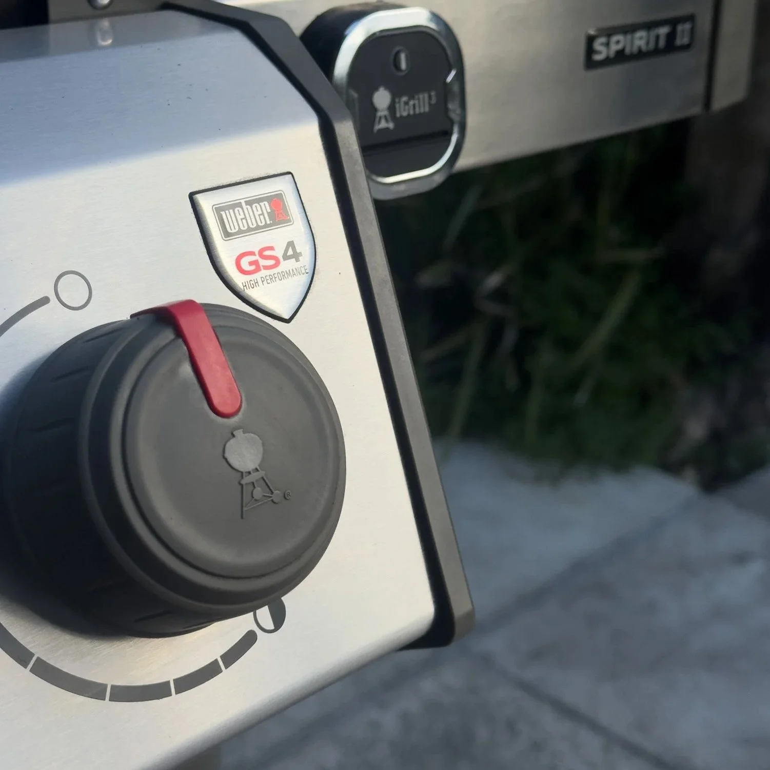 Close-up of a Weber Spirit II gas grill control panel showing a black temperature control knob with a red indicator, a GS4 high performance badge, and an ignition button.