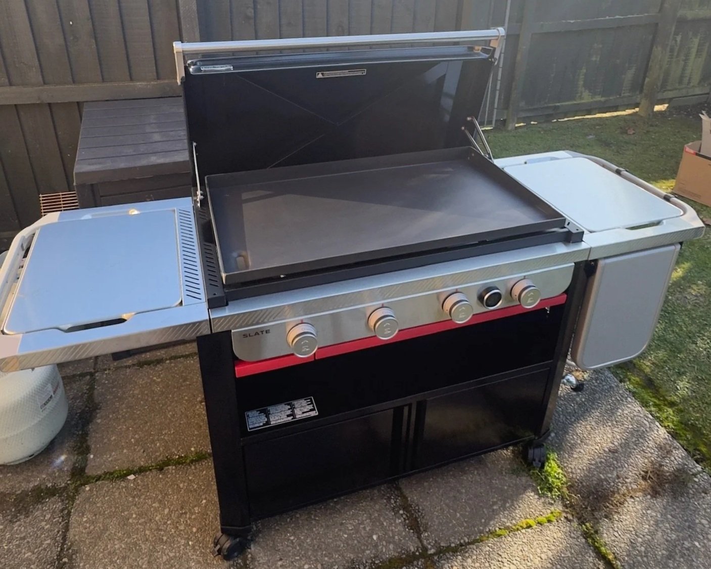 Outdoor stainless steel commercial griddle with four control knobs on a black grill base, accompanied by a side burner, on a paved patio with a wooden fence and grass in the background.