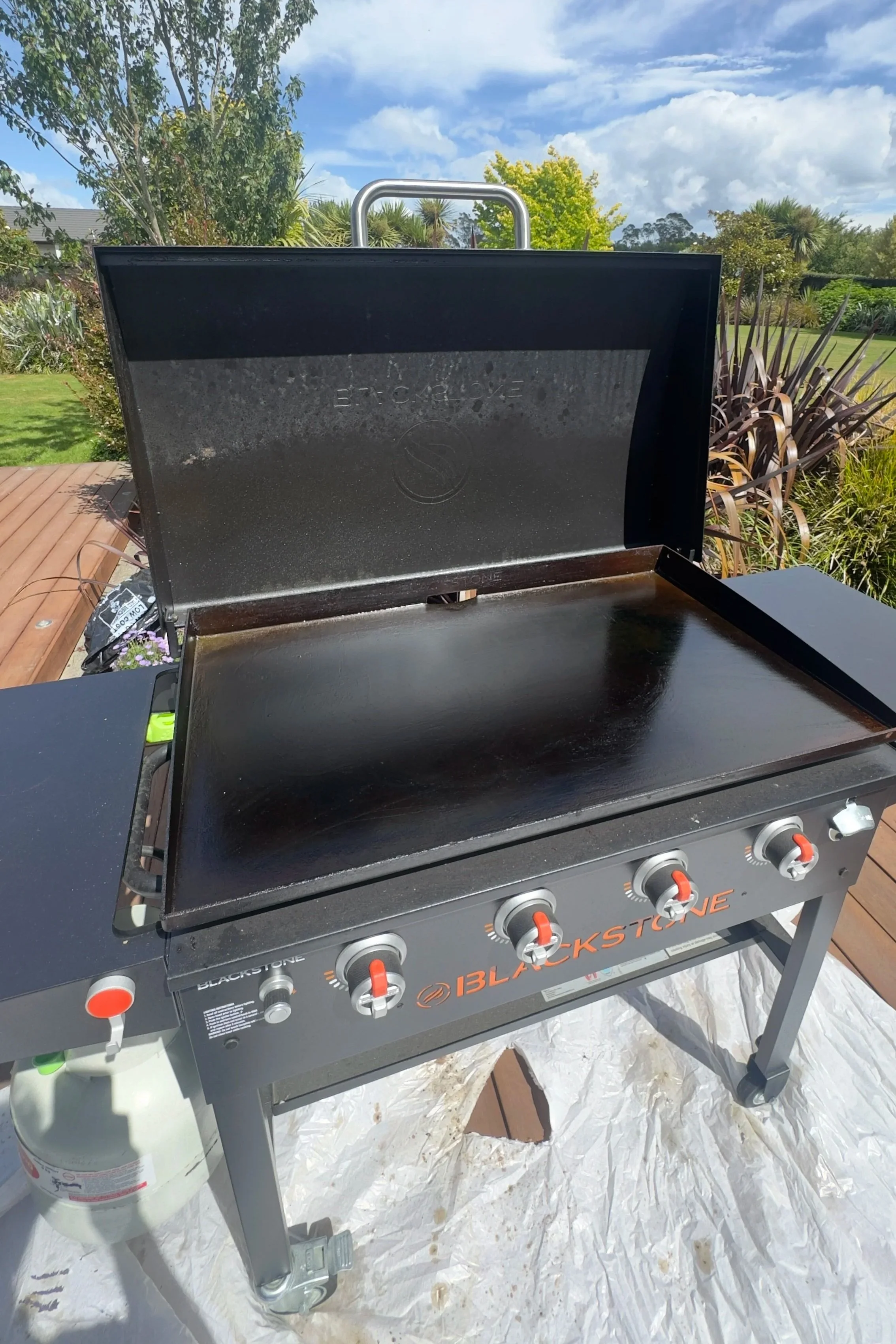 Blackstone outdoor gas grill with an open lid, four control knobs, and a propane tank underneath, situated on a wooden deck with a garden and trees in the background, under a blue sky with clouds.
