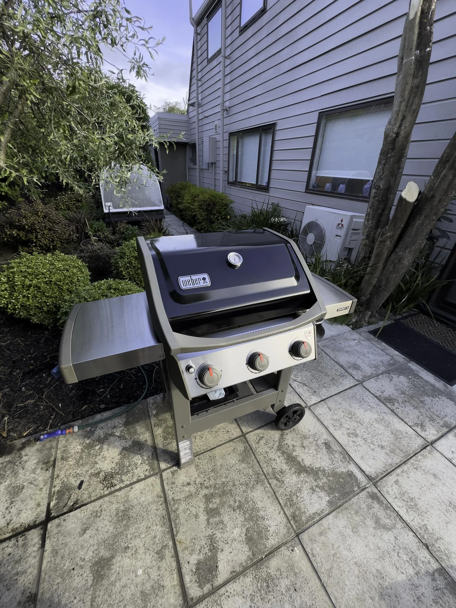 Silver gas grill on patio near house with gray siding, tree, small bushes, and air conditioning unit in backyard.