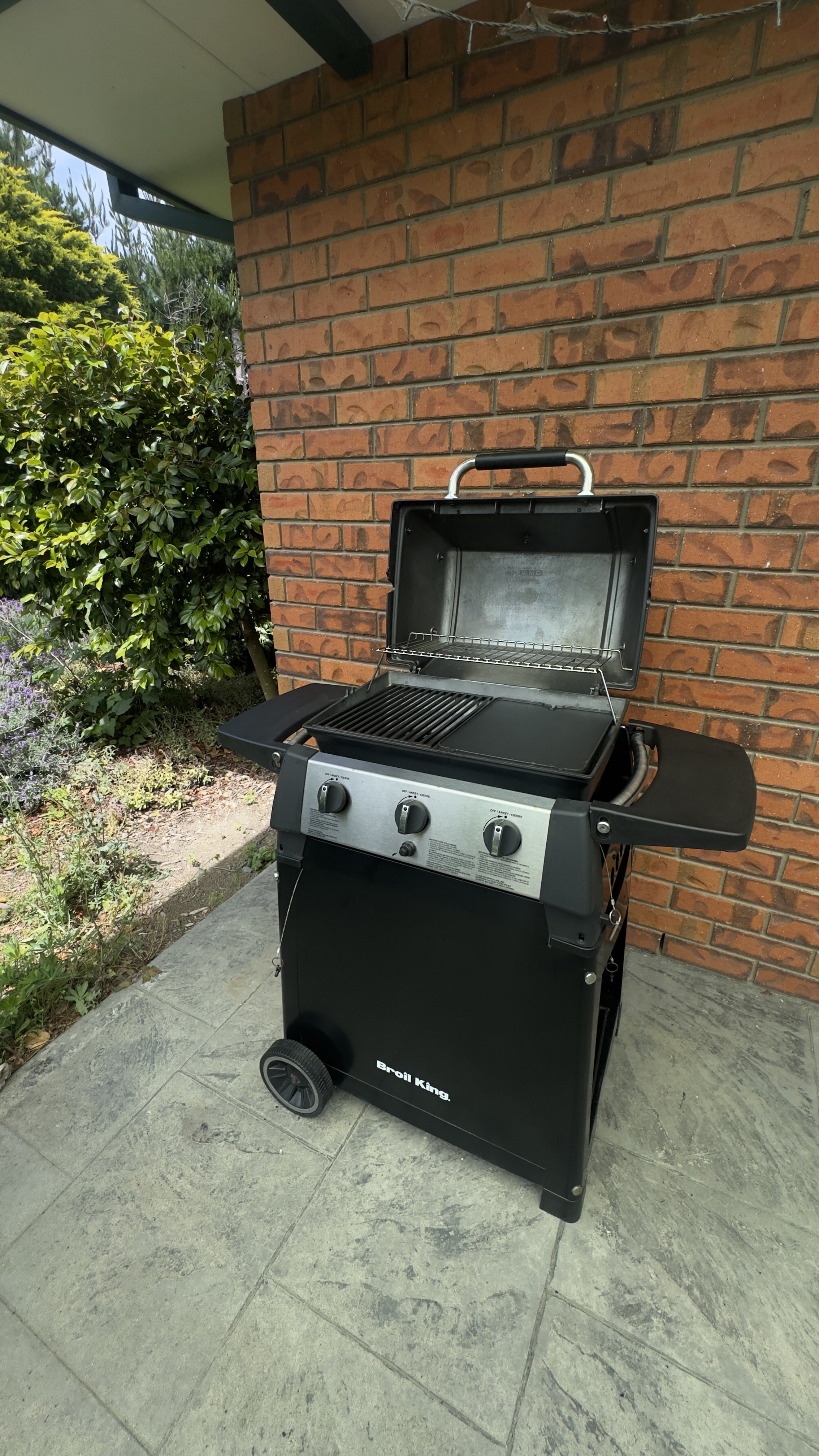 A black Brosil King gas grill with its lid open, showing grilling and warming racks, positioned on a concrete patio beside a brick wall and some greenery.