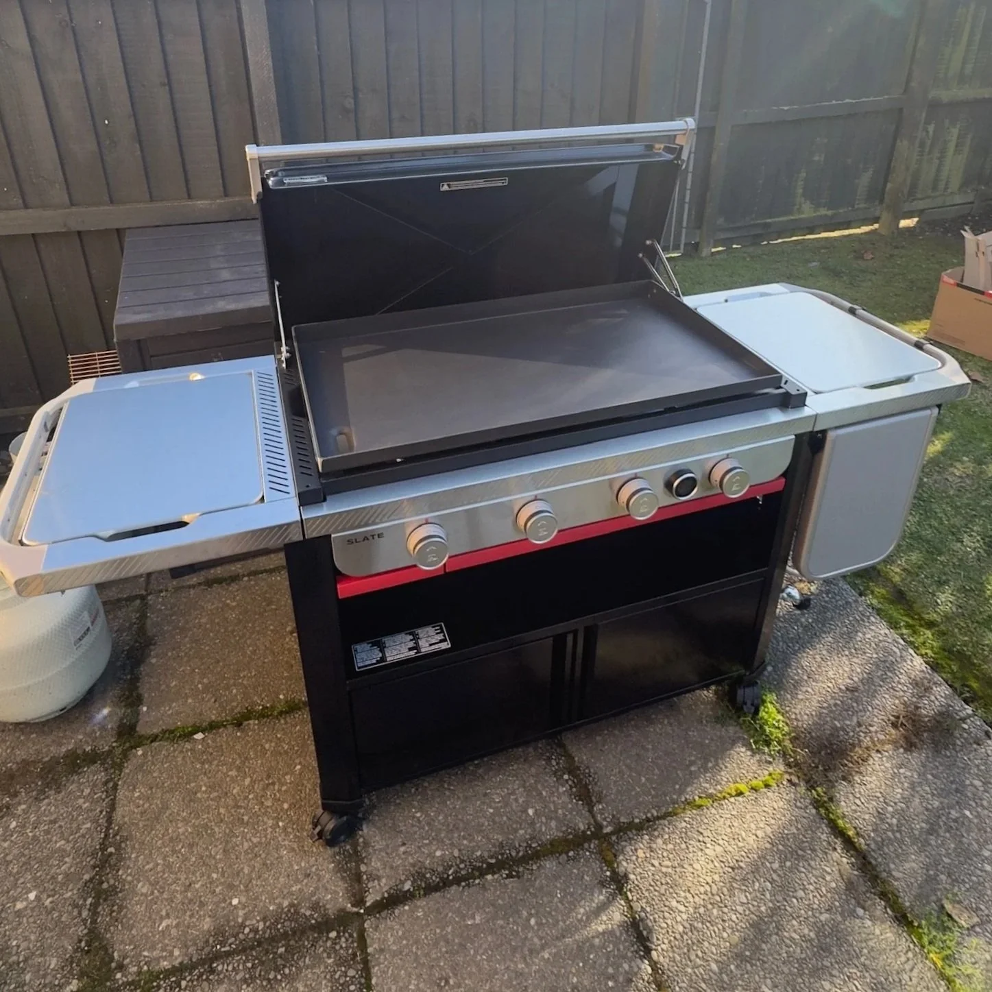 Outdoor propane gas grill with a black and stainless steel finish, multiple control knobs, attached side shelves, and a closed hood, situated on a paved patio area with a wooden fence in the background.