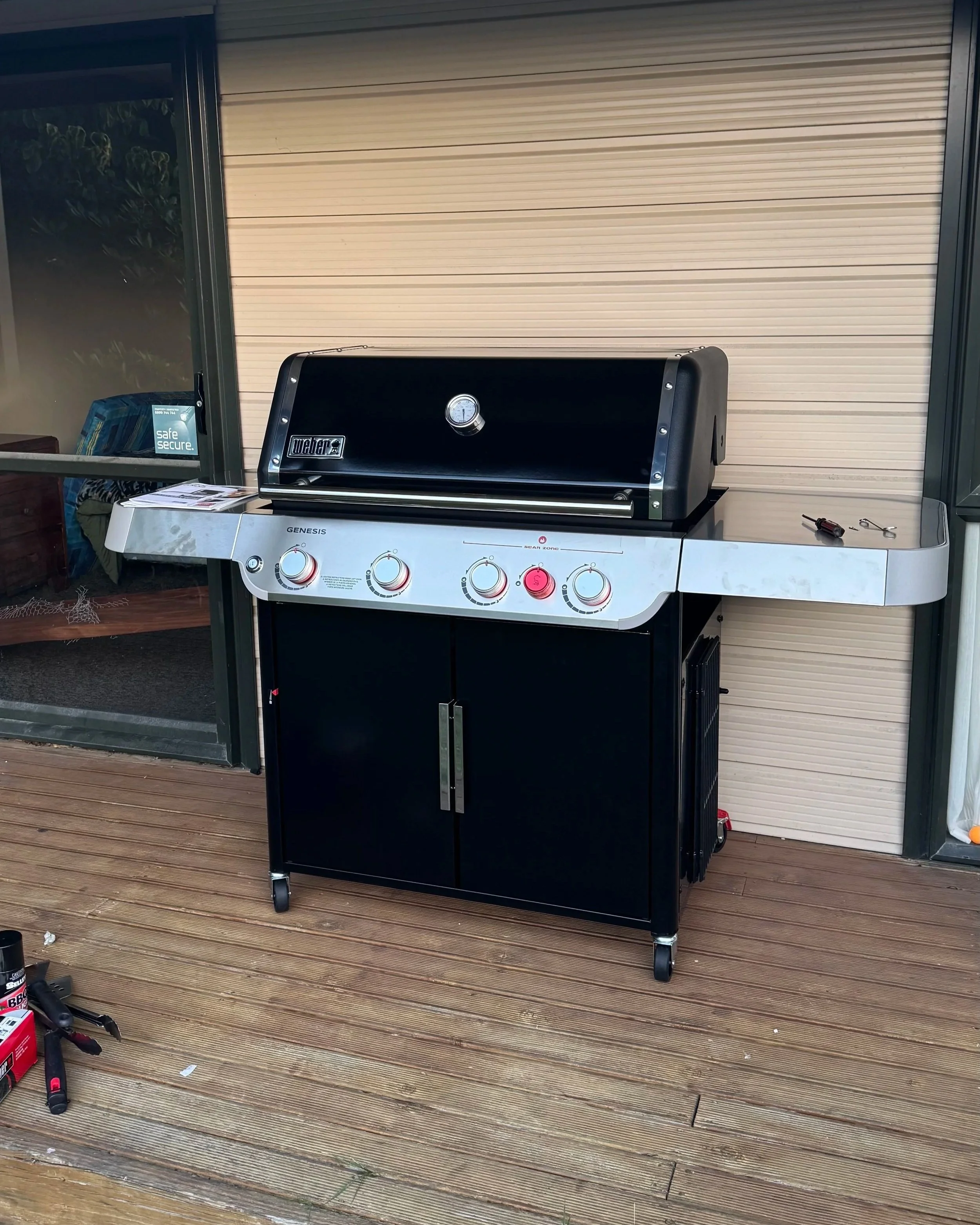 A black and silver outdoor gas grill on wheels, placed on a wooden deck, with tools on the side and a small booklet.
