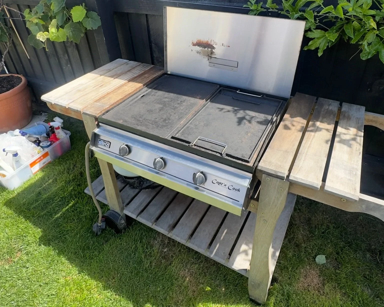 A portable gas grill with a metal lid and control knobs on a wooden outdoor table, with gardening supplies in a plastic container nearby.