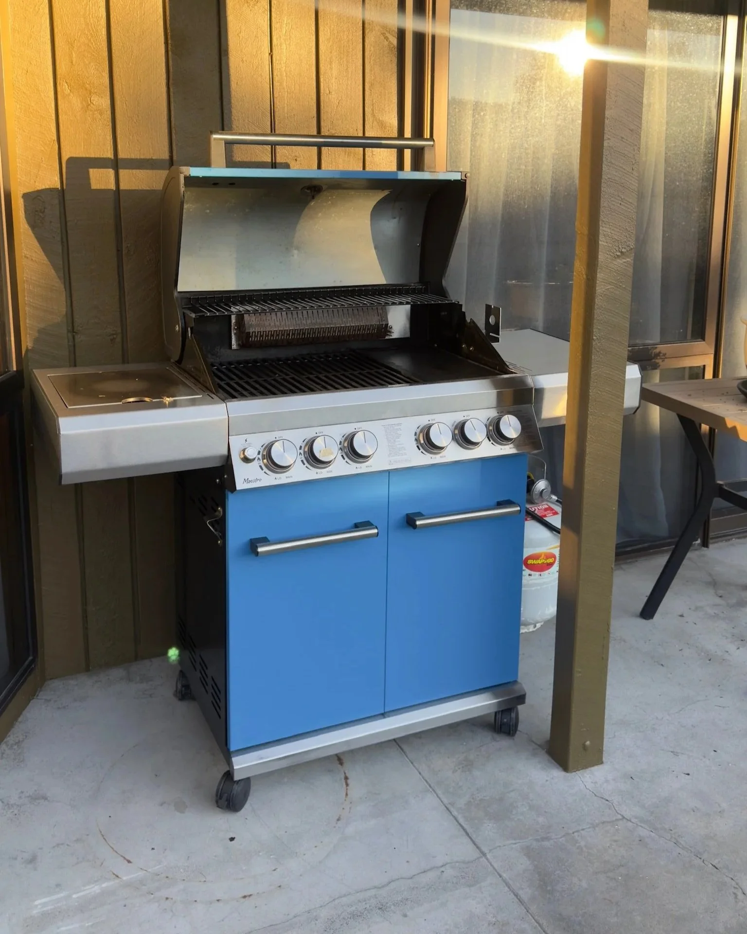 A blue and stainless steel outdoor gas grill with four control knobs, positioned on a concrete porch next to a wooden post and a sliding glass door, with sunlight shining from the upper right.