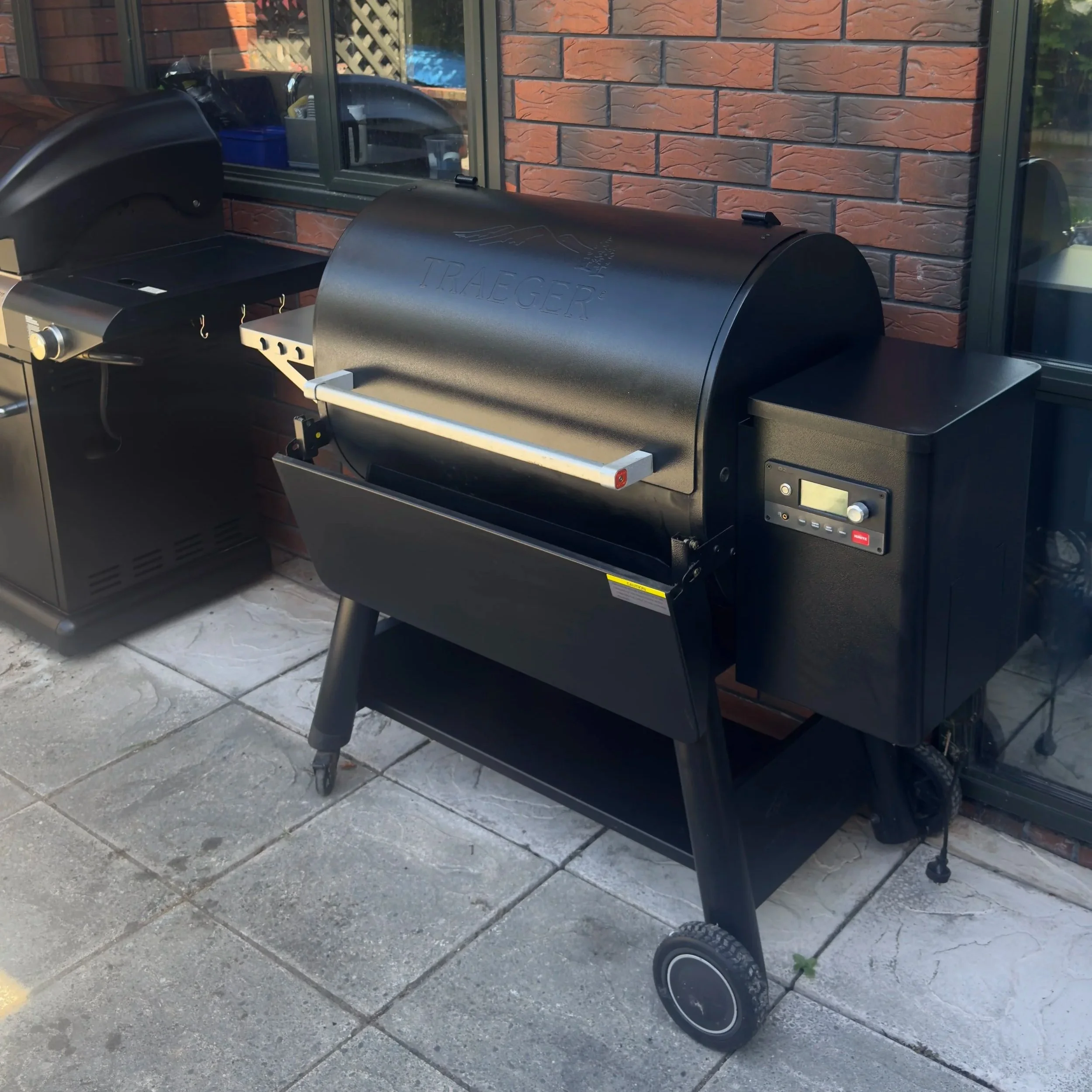 Black Traeger pellet grill barbecue on a patio with brick wall and windows in the background.