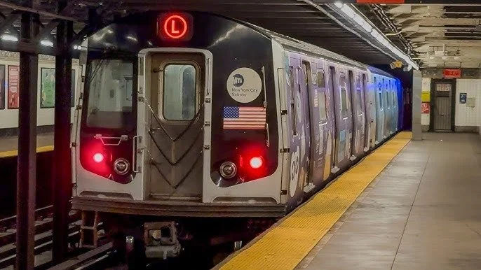 New York City subway train with an L line indicator, arriving at an underground station platform.