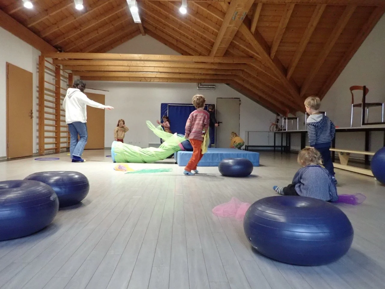 Kinder spielen in einem Raum mit Holzbalkendecke, einige sitzen auf blauen Bällen, andere spielen mit einer grünen Tuchrolle auf einer Matratze.
