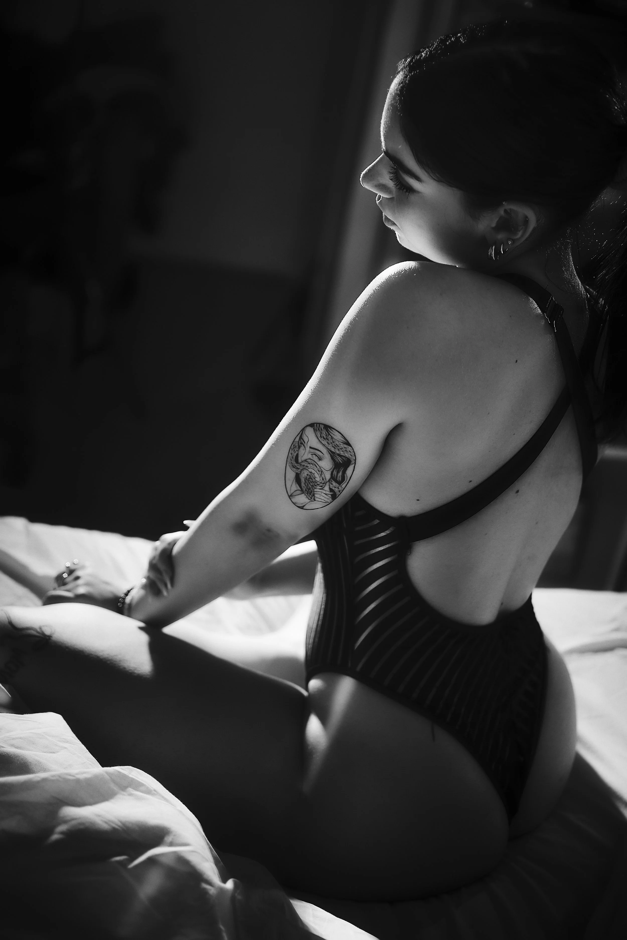 Photo pour séance boudoir, portrait intime. Une jeune femme avec un tatouage sur le bras, assise sur un lit, en train de se coiffer ou de se préparer, dans un éclairage en noir et blanc.