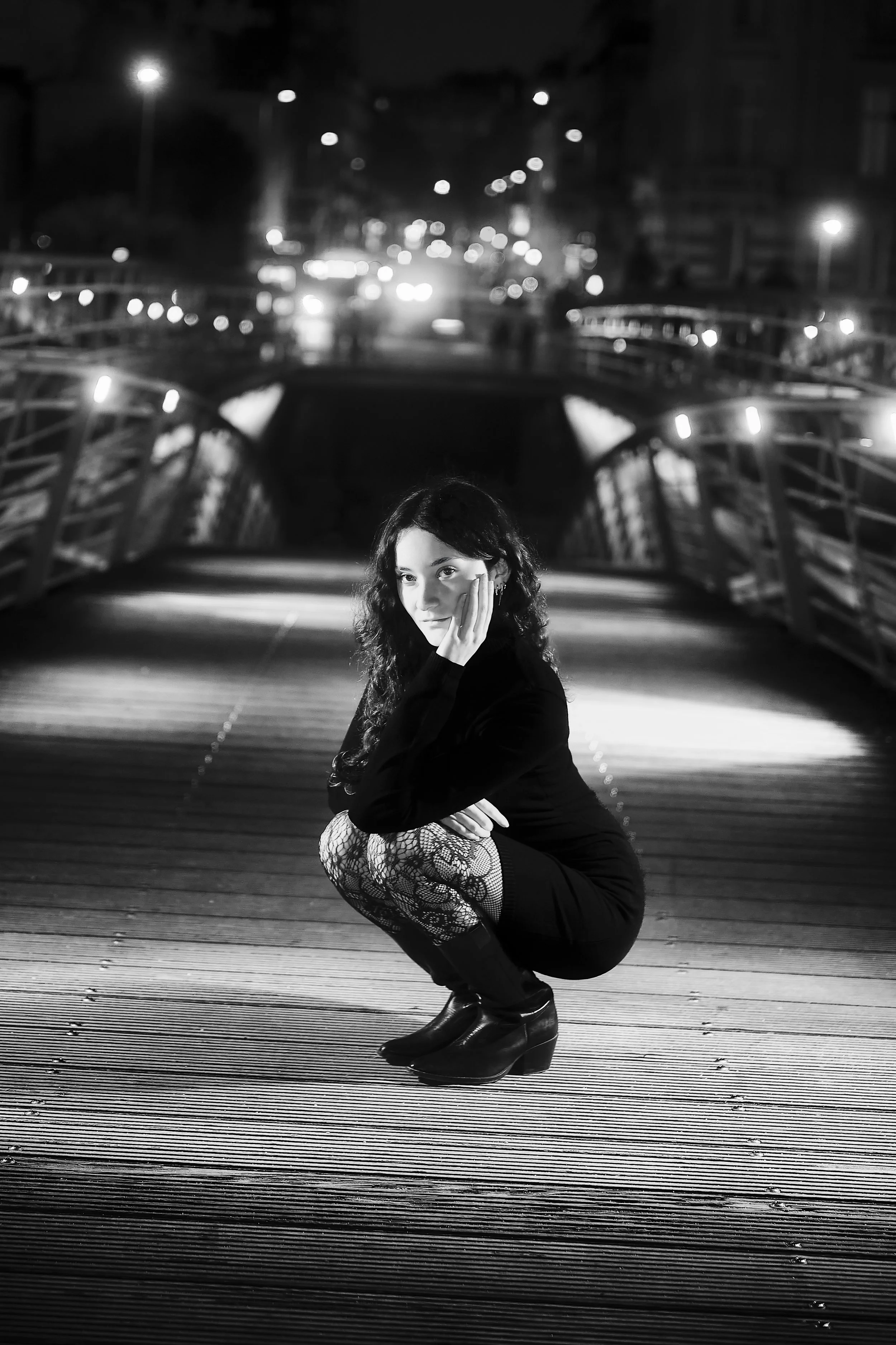 Photo pour séance Portrait Intimiste. Une femme en vêtements noirs, portant des collants en dentelle et des bottines, est assise en position accroupie sur la passerelle Léopold-Sédar-Senghor la nuit dans Paris, avec un fond urbain flou et éclairé