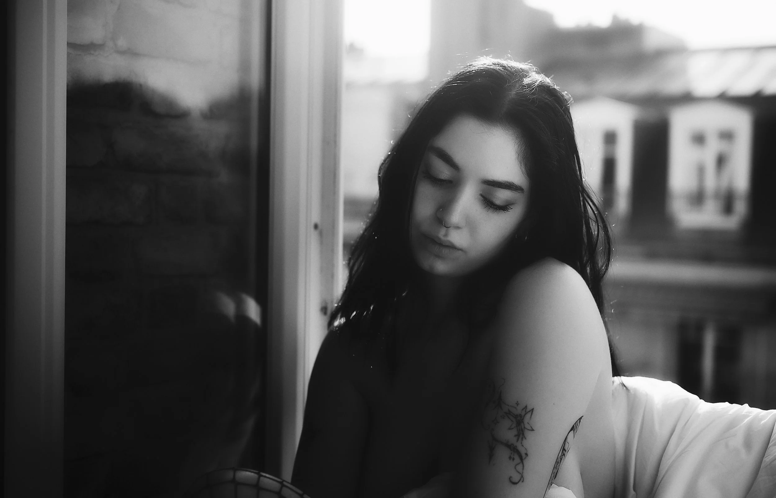 Photo en noir et blanc pour séance boudoir. Jeune femme aux cheveux noirs, yeux fermés, avec un tatouage sur le bras, assise près d'une fenêtre, dans une pièce en style minimaliste.