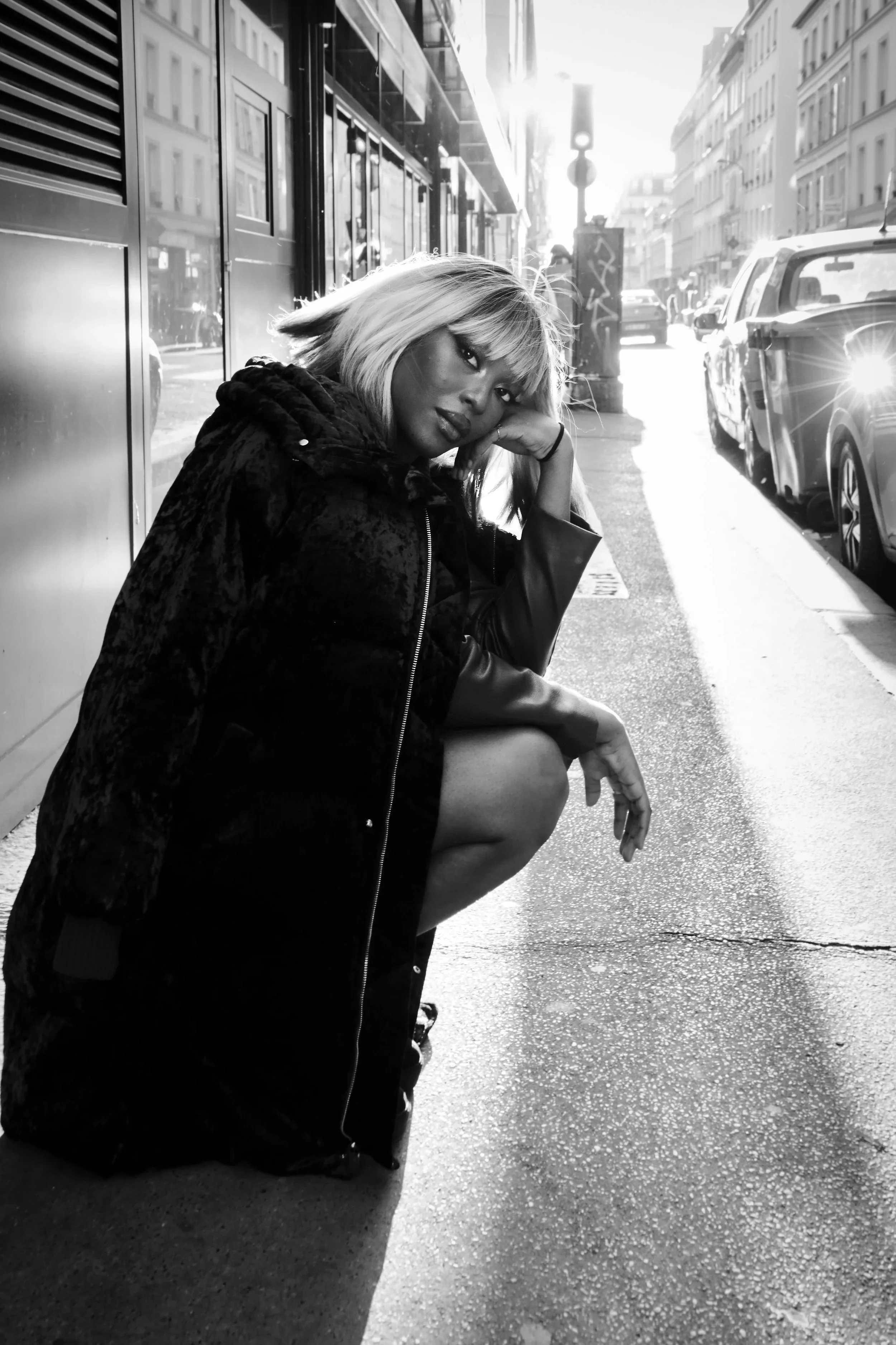 Photo pour séance Portrait Intimiste. Femme assise au sol sur un trottoir, portant un manteau foncé, dans une rue urbaine avec des voitures et des bâtiments en arrière-plan, en noir et blanc.