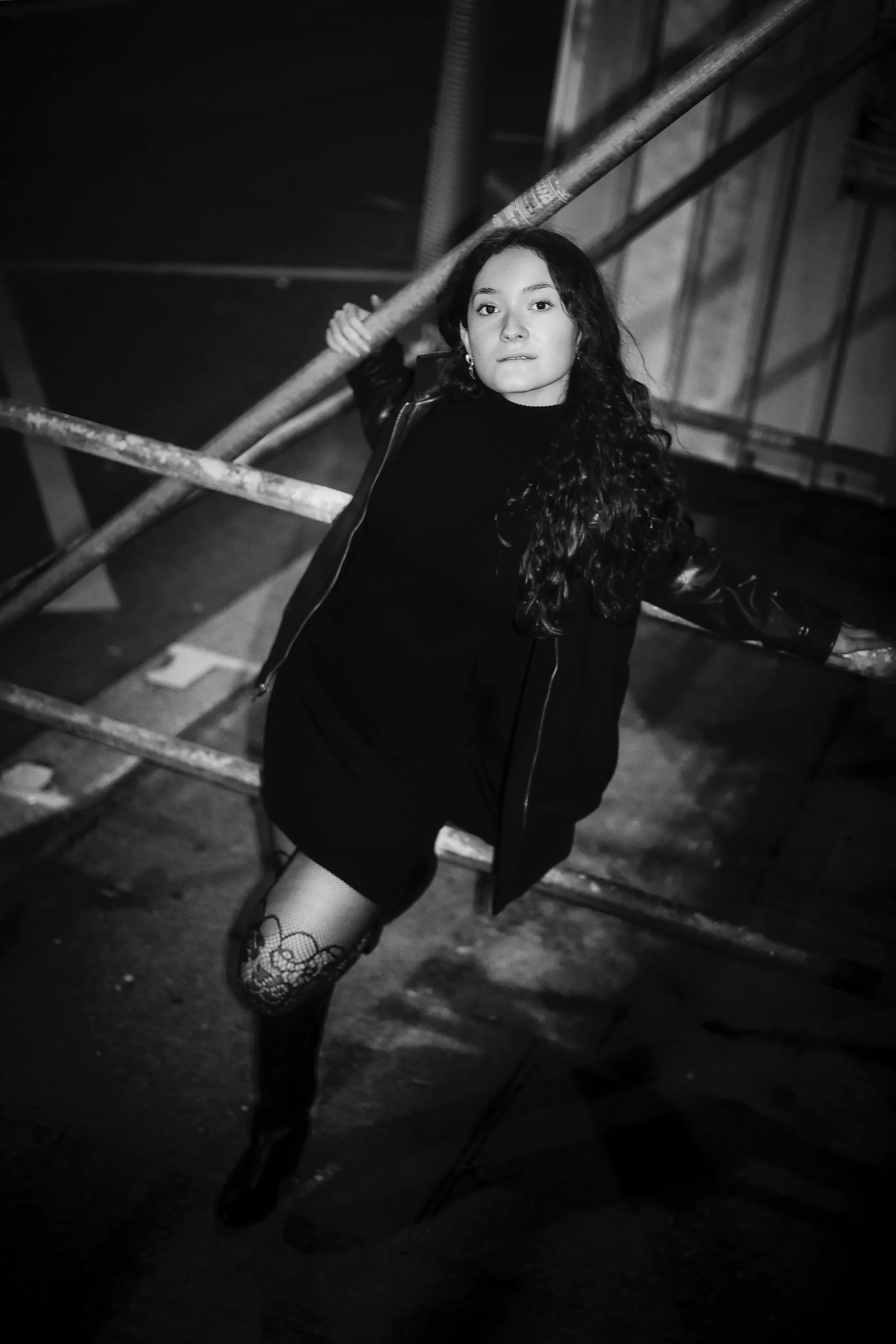 Photo pour séance Portrait Intimiste. Jeune femme aux cheveux bouclés, portant un vêtement noir, posant contre une barrière métallique dans un environnement urbain sombre, en noir et blanc.