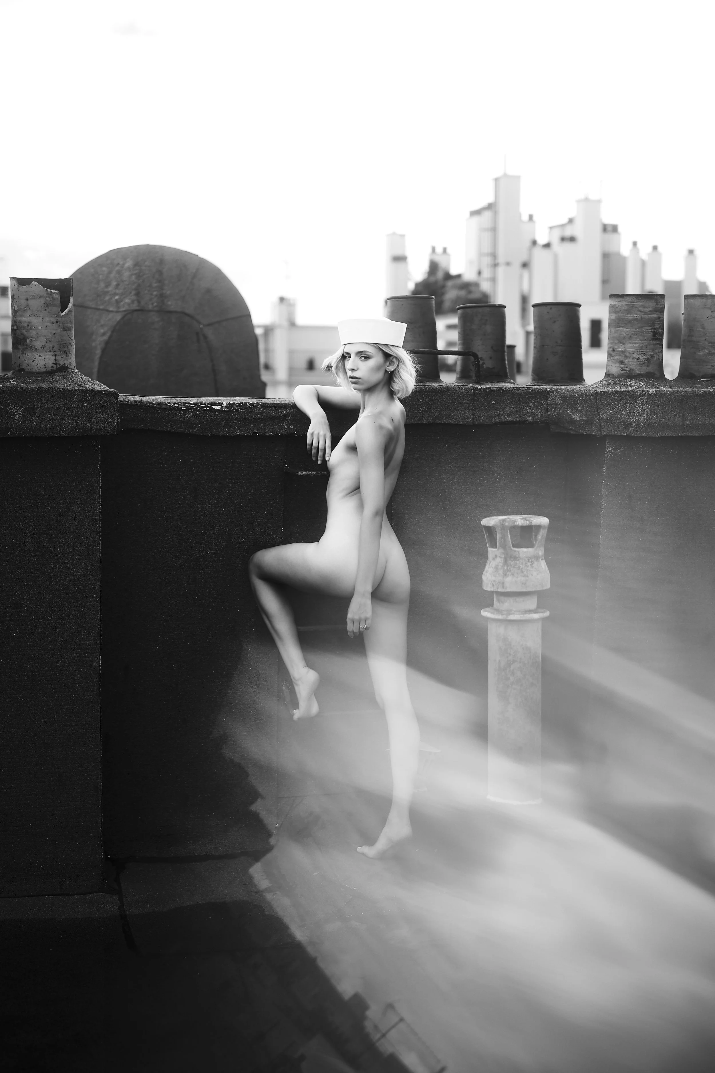 Photo pour séance Nue. Une femme nue portant une coiffe blanche, debout sur un toit avec une vue urbaine en arrière-plan, dans une photo en noir et blanc.