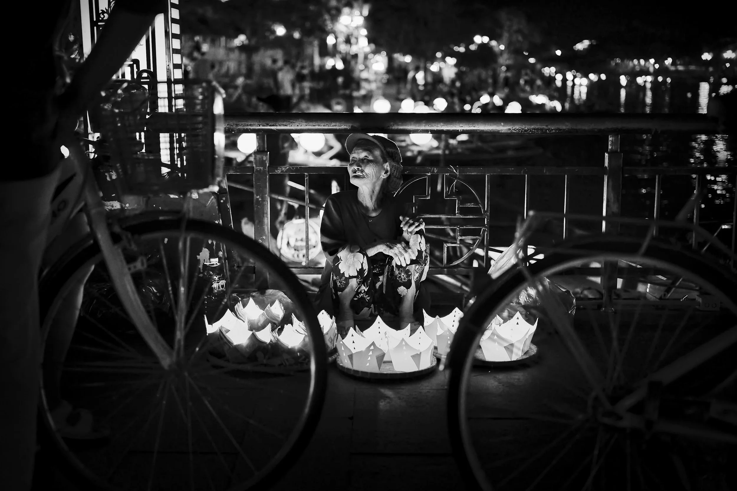 Une femme assise derrière une table décorée de lanternes en papier illuminées, entourée de bicyclettes, dans un environnement nocturne avec des lumières en arrière-plan.