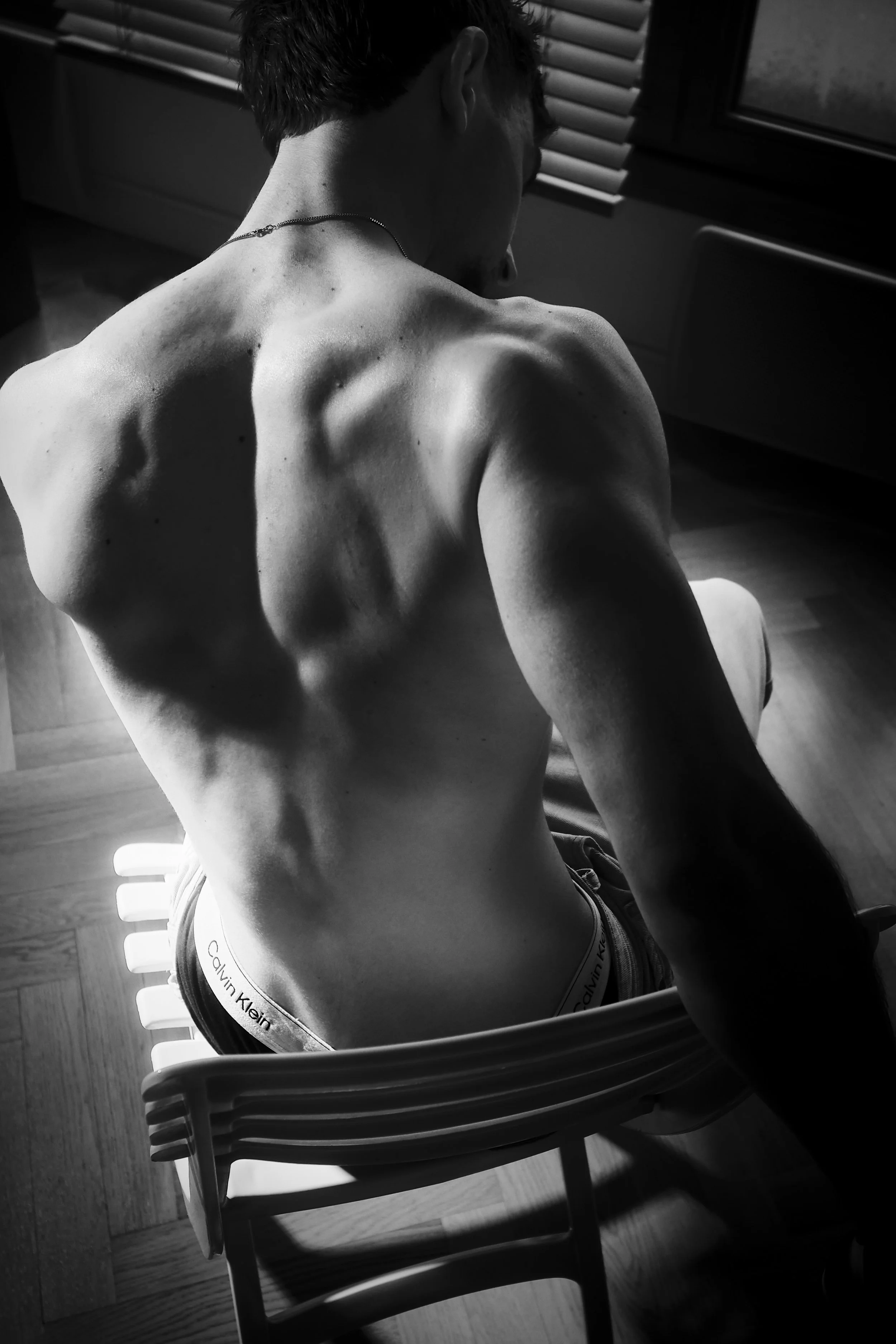 ~ Lucas ~ 
Photo en noir et blanc d'un jeune homme nu vu de dos, assis sur une chaise, avec la tête légèrement inclinée. Il porte un collier et un sous-vêtement Calvin Klein. Photographie artistique fine art. Séance photo boudoir Paris. Photographe i