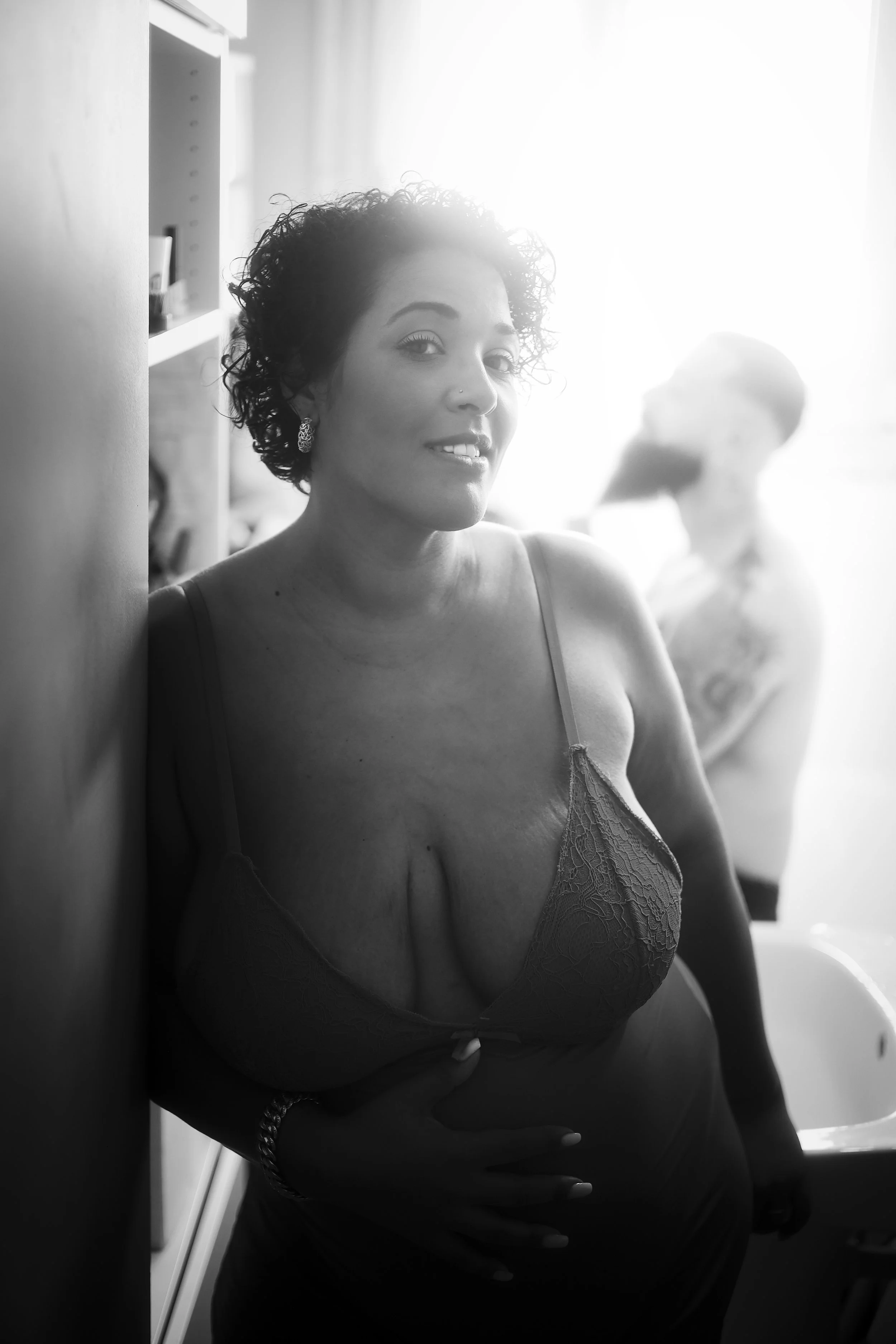 Photo pour séance portrait intimiste. Une femme avec des cheveux bouclés courts, portant un haut en dentelle et des boucles d'oreilles, sourit légèrement devant une vitre ou une lumière intense en arrière-plan.