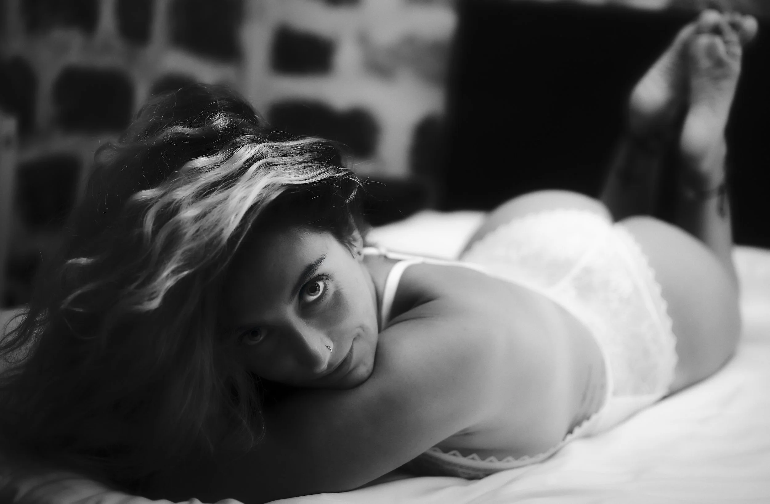 Photo pour séance Boudoir. Une femme allongée sur un lit, regardant la caméra, en lingerie blanche, avec des cheveux longs et ondulés, dans une pièce avec un mur en pierre en arrière-plan, en noir et blanc.