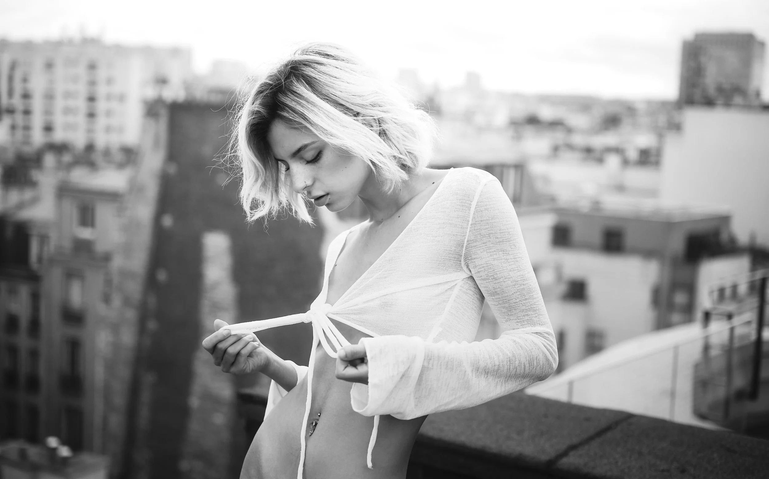 Photo pour séance Portrait intimiste sur les toits de Paris. .Jeune femme aux cheveux courts et bouclés, portant un haut transparent blanc qu'elle ajuste en le tirant par les liens, se tenant sur un toit avec une vue urbaine floue en arrière-plan.