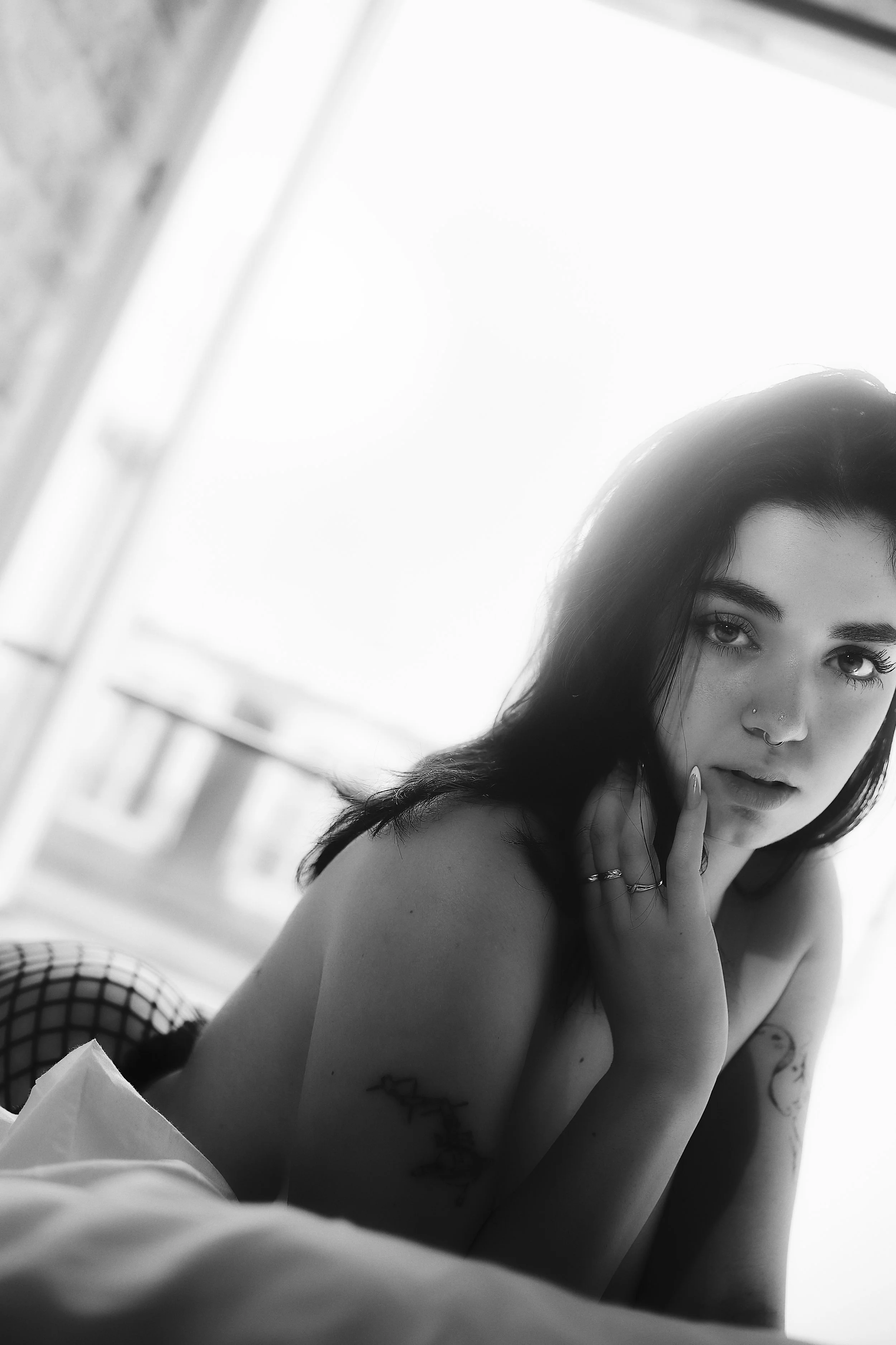 Photo pour séance Portrait Boudoir. Photo en noir et blanc d'une femme aux cheveux foncés, avec un piercing au nez, posant avec une main près du visage, dans une pièce lumineuse.