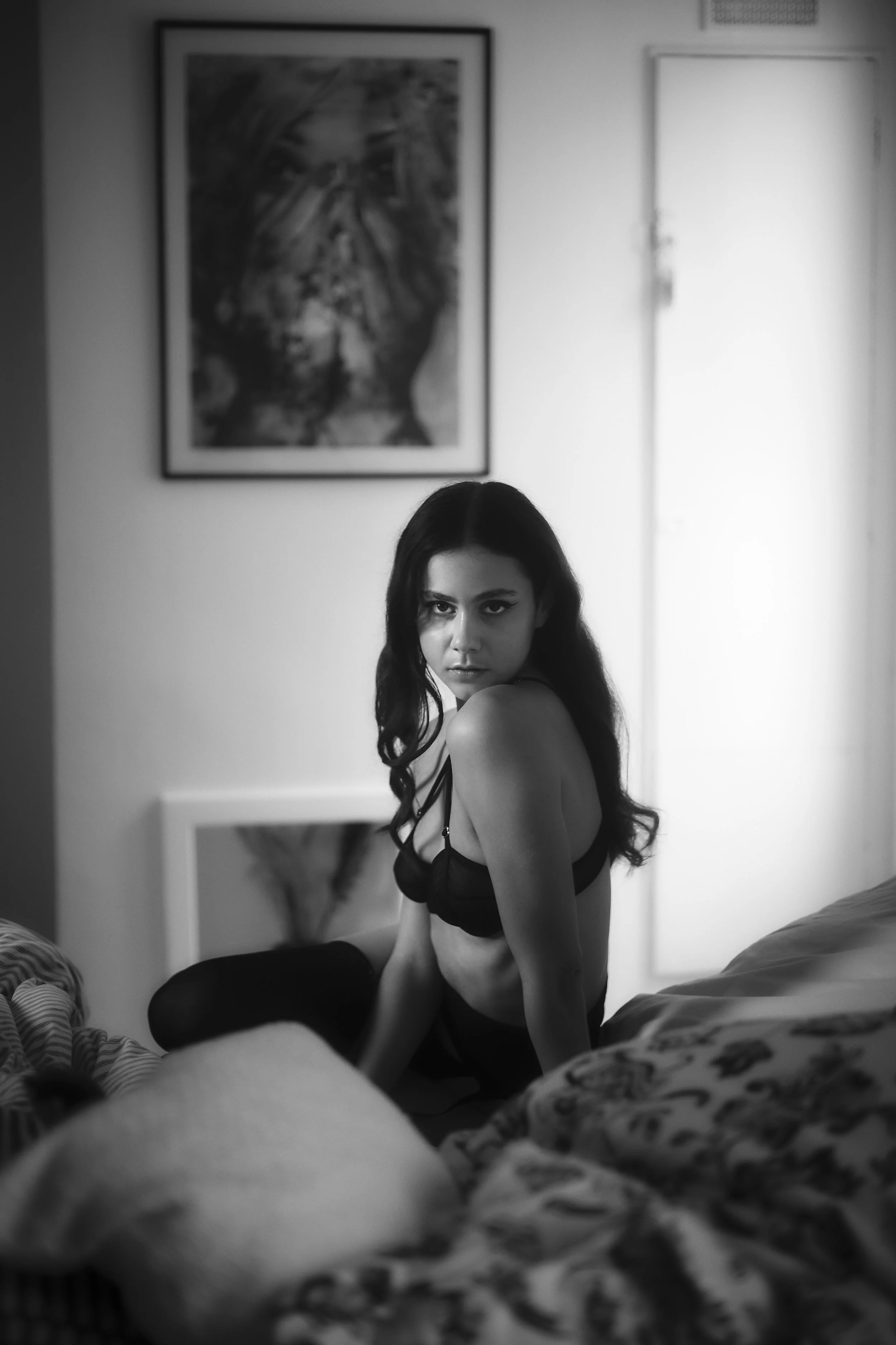 Portrait en noir et blanc d'une femme avec des cheveux longs portant un soutien-gorge noir, assise sur un lit dans une pièce, regardant fixement l'objectif, en noir et blanc.