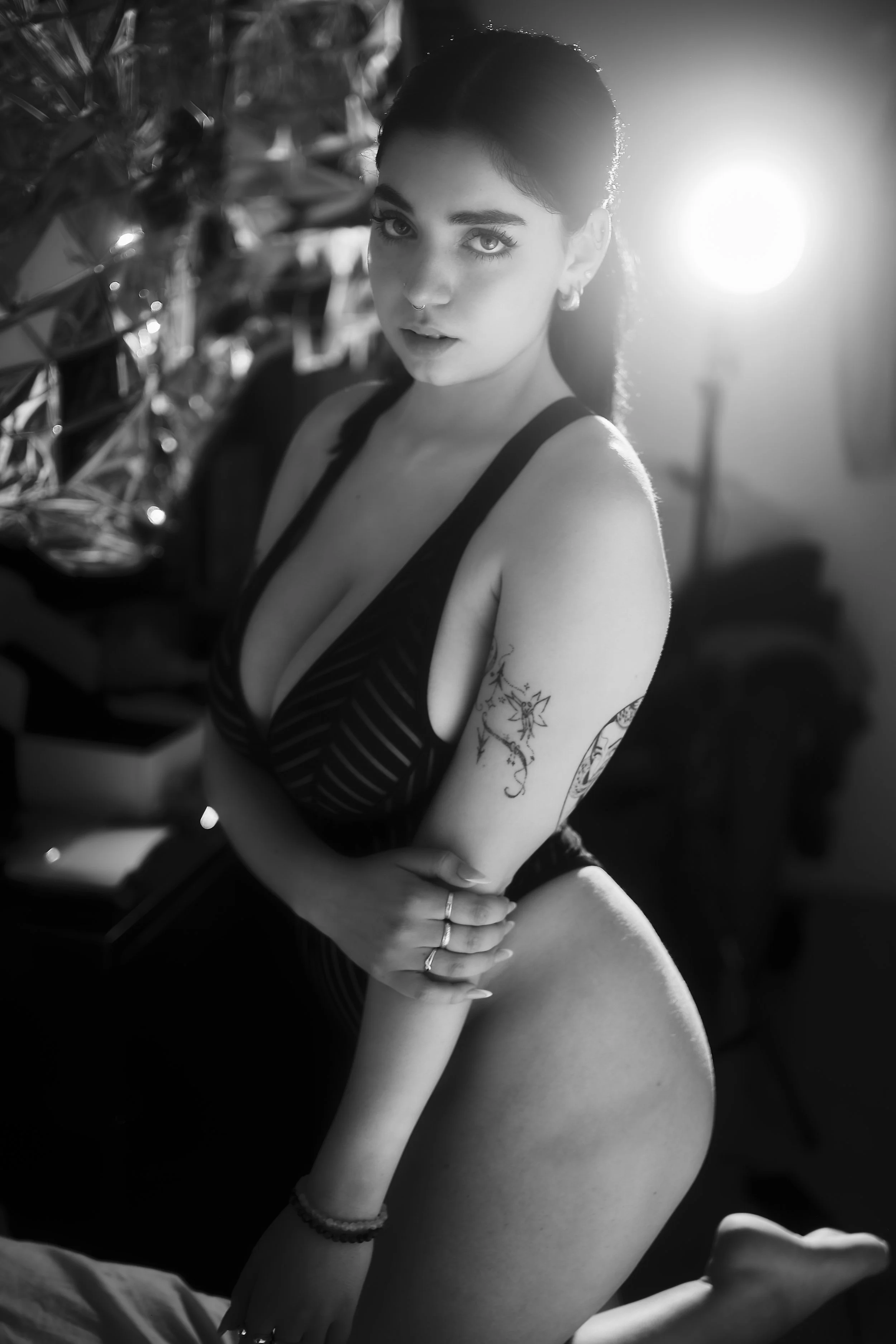 Photo pour séance portrait charme. Une femme jeune avec des tatouages, portant un haut à rayures, posant de profil avec un regard vers l'objectif, dans un environnement avec une forte lumière derrière elle, en noir et blanc.