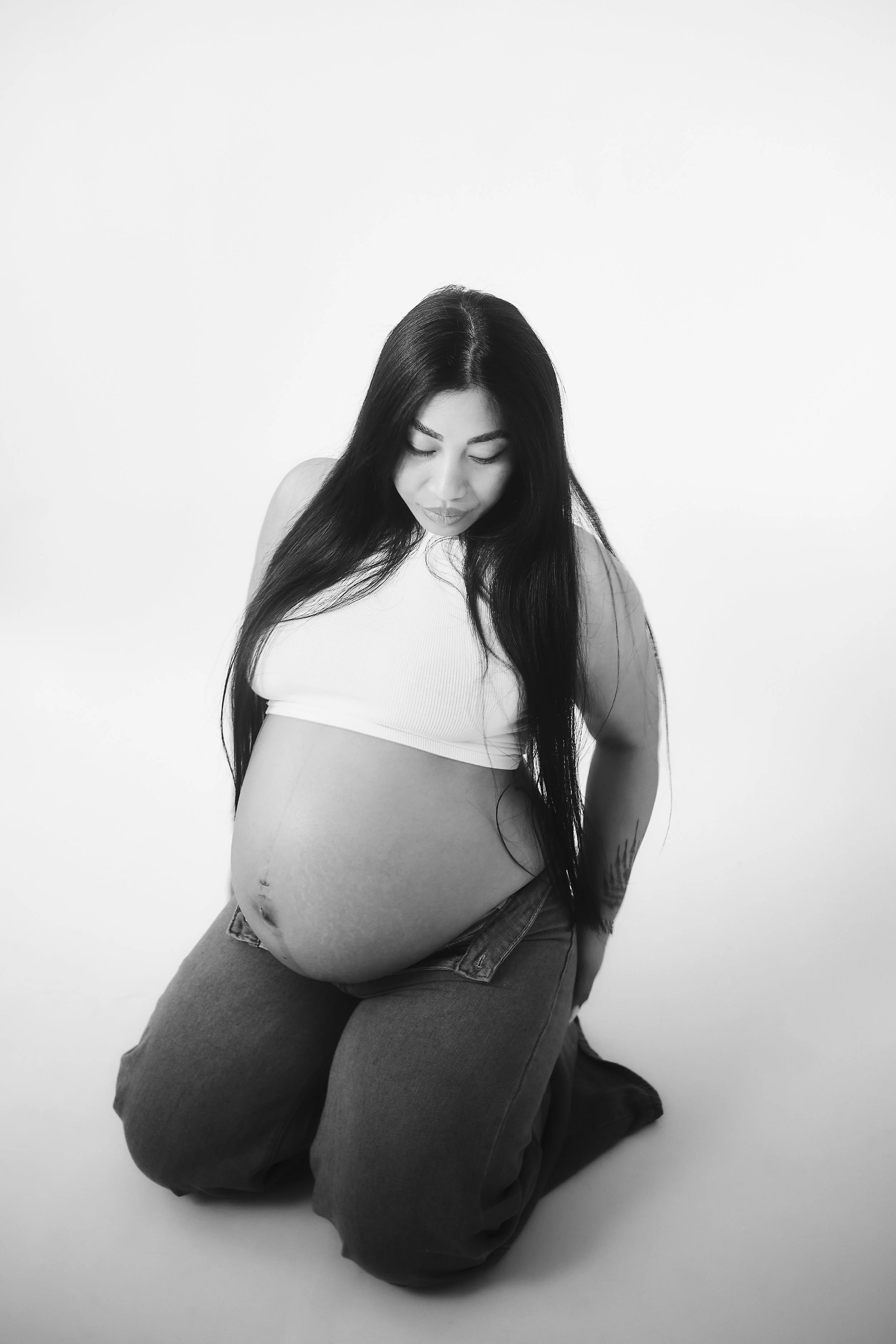 Photo pour séance Portrait Intimiste. Une femme enceinte avec de longs cheveux noirs, portant un haut blanc et un pantalon foncé, est agenouillée sur un sol blanc, regardant son ventre.