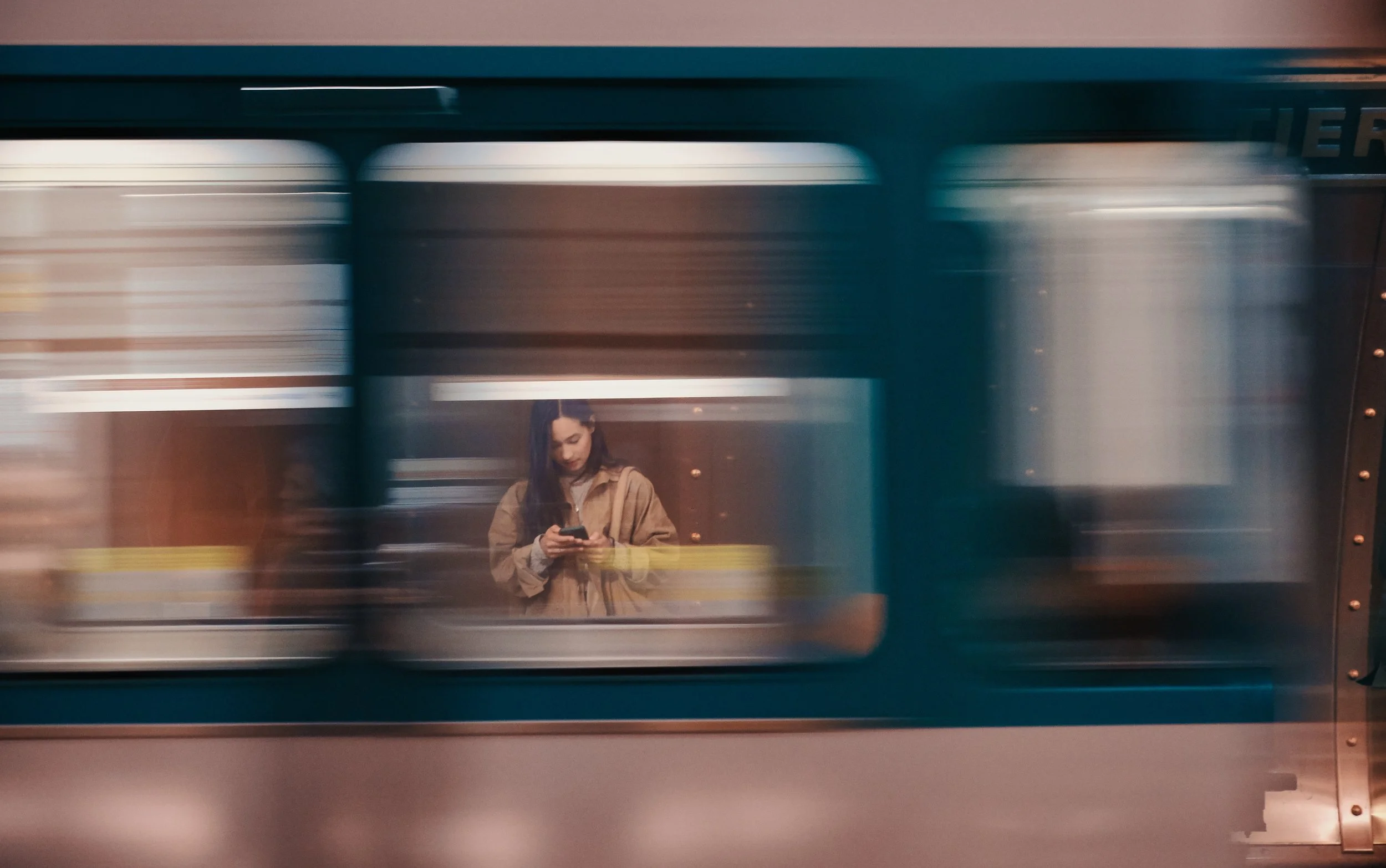 Une jeune femme avec une veste beige se trouve dans une station de métro, regardant son téléphone, à travers une vitre de train en mouvement.