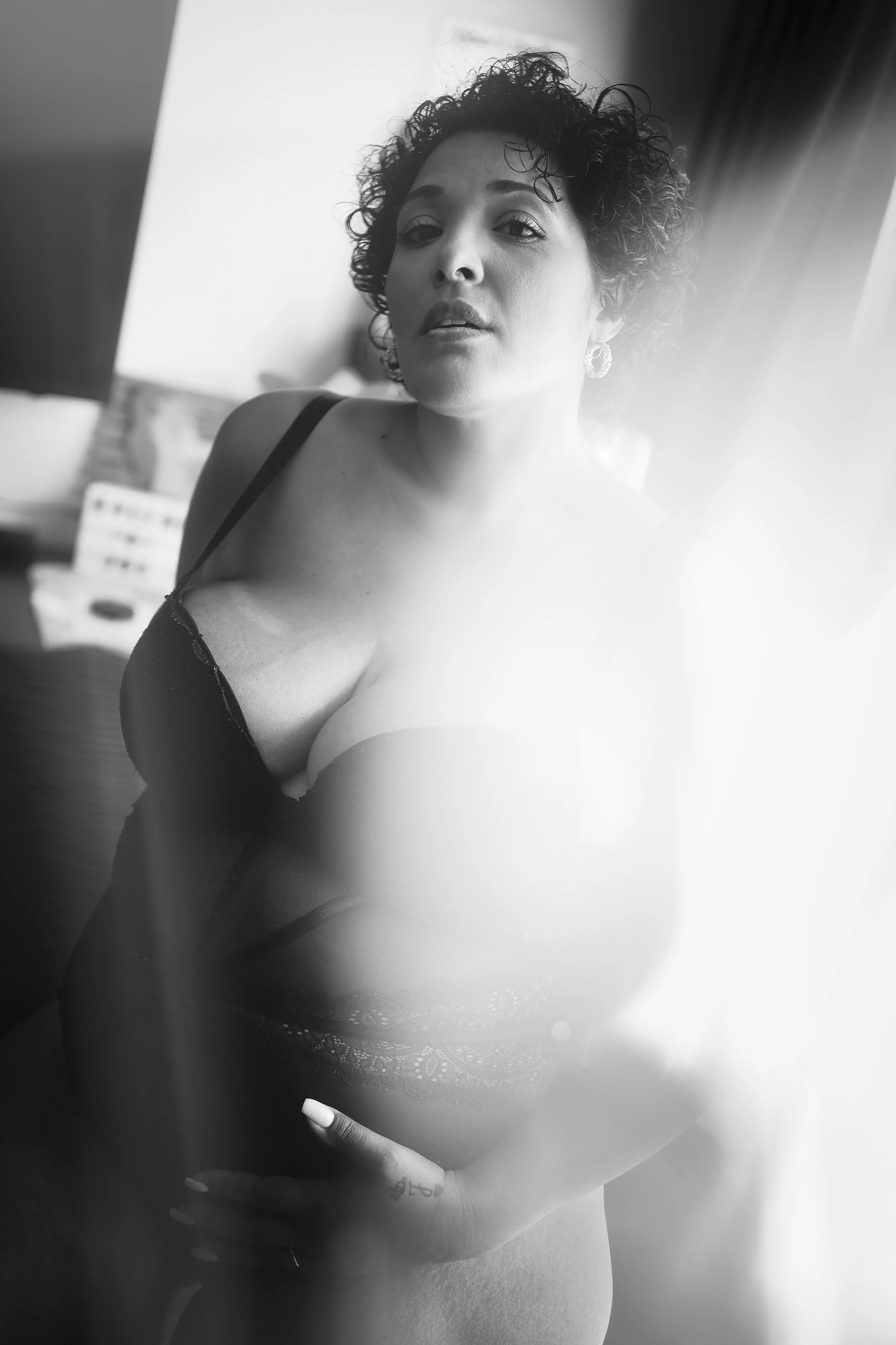 Photo pour séance boudoir. Une femme en lingerie noire, tenant son ventre, avec un fond flou, en noir et blanc.