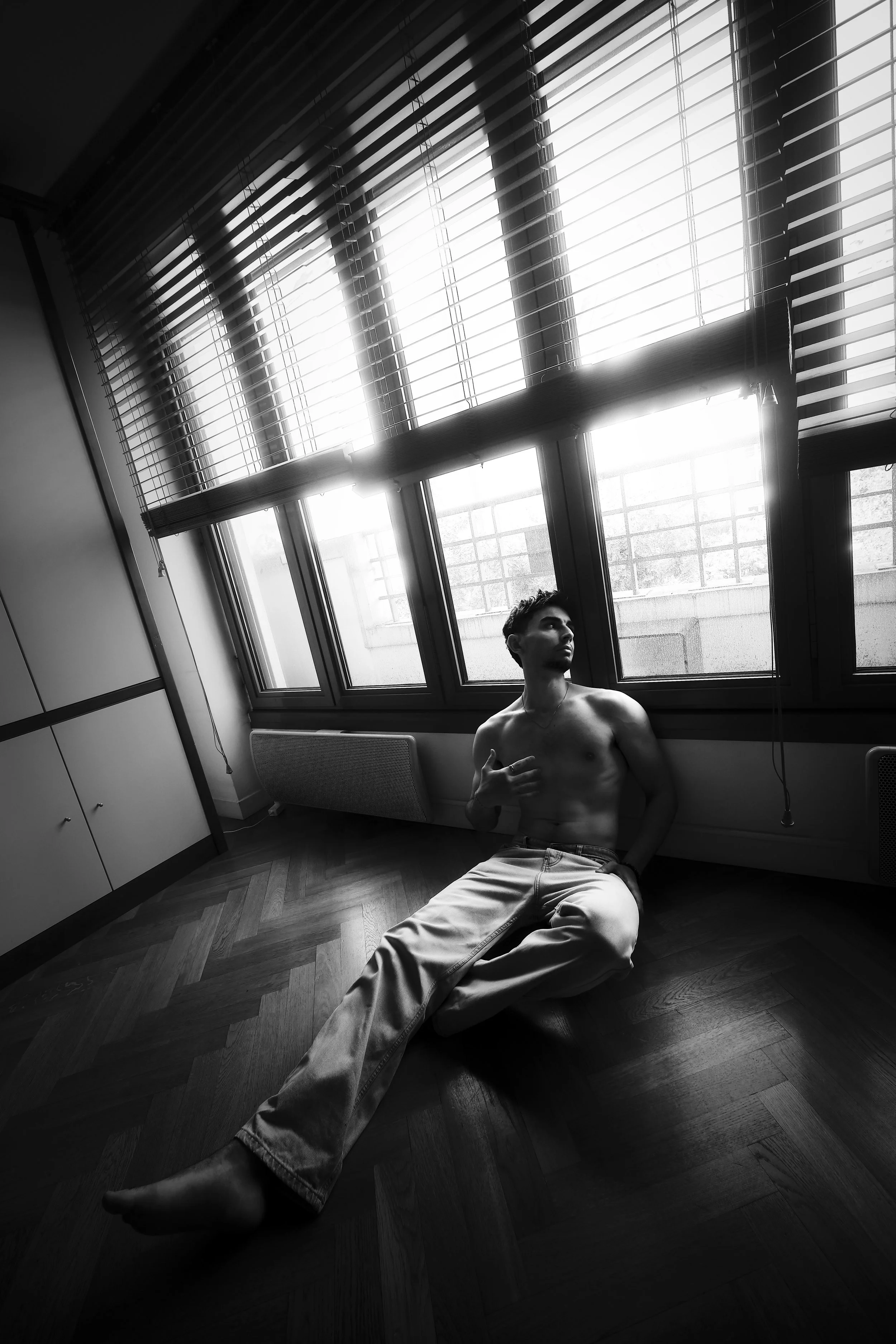 ~ Lucas ~ 
Portrait d'un jeune homme torse nu assis sur le sol d'une pièce, près d'une grande fenêtre avec des volets, dans une photographie en noir et blanc. Photographie artistique fine art. Séance photo boudoir Paris. Photographe intimiste Paris.