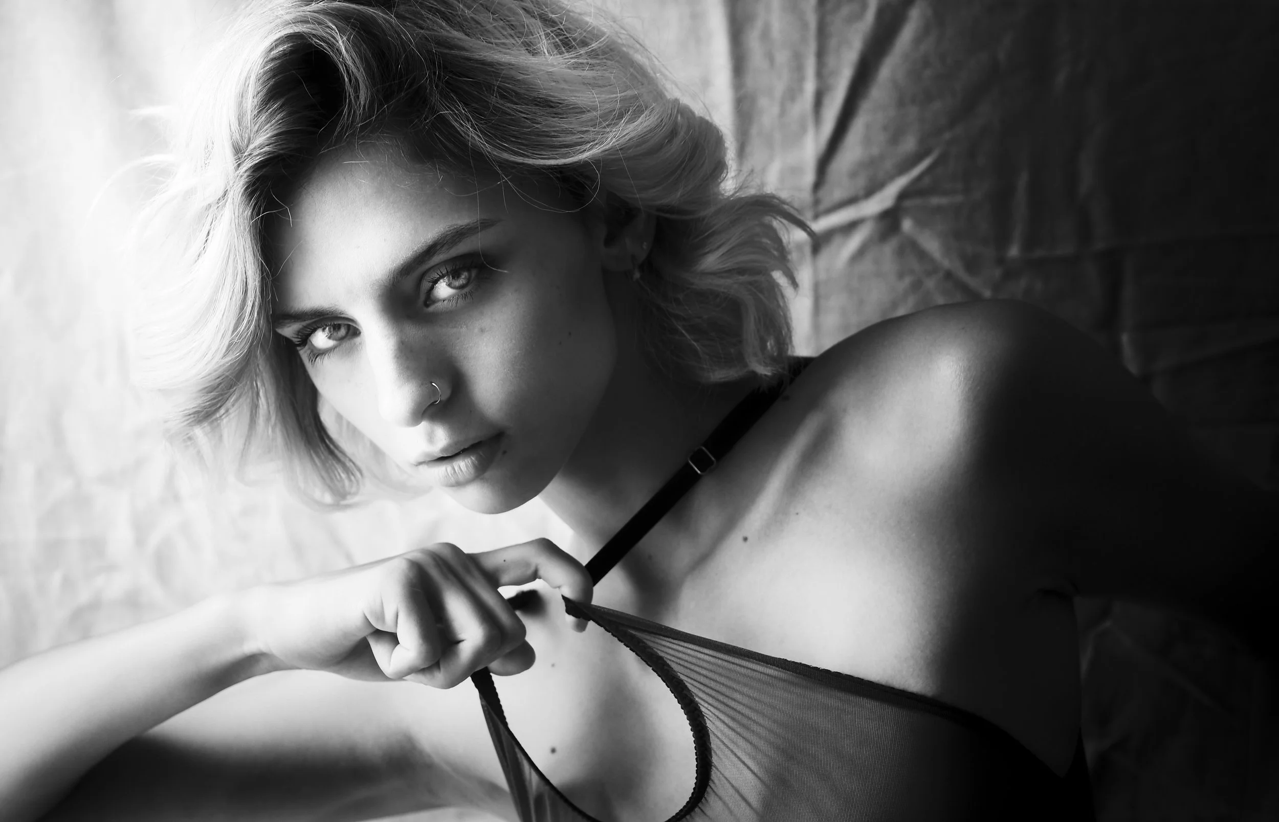 Photo en noir et blanc pour séance portrait boudoir. Portrait en noir et blanc d'une jeune femme aux cheveux blonds, portant un haut transparent, regardant intensément vers l'objectif.