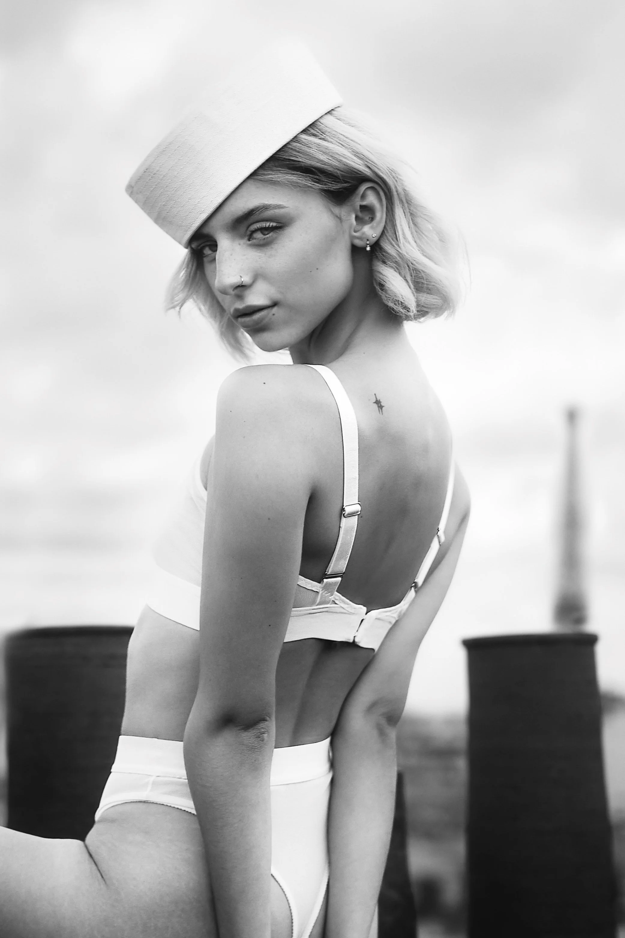 Photo noir et blanc pour une séance portrait boudoir, portrait intimiste. Une femme avec un chapeau blanc, des cheveux blonds, portant un haut blanc à bretelles. Posant sur les toits de paris.