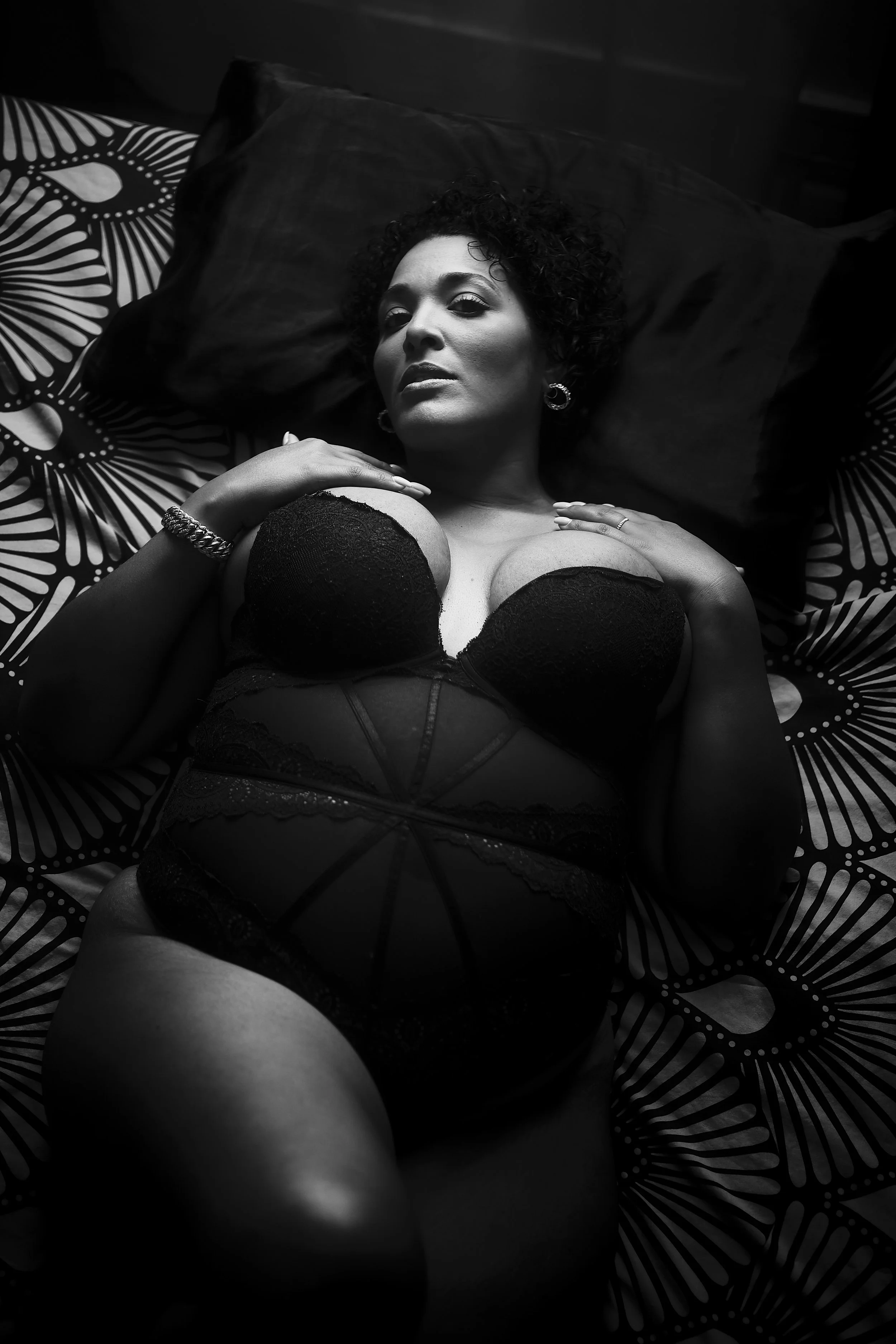 ~ Kalinda ~
Portrait en noir et blanc d'une femme allongée sur un lit, vêtue de lingerie noire, regardant vers l'appareil, avec des motifs floral sur la parure de lit.