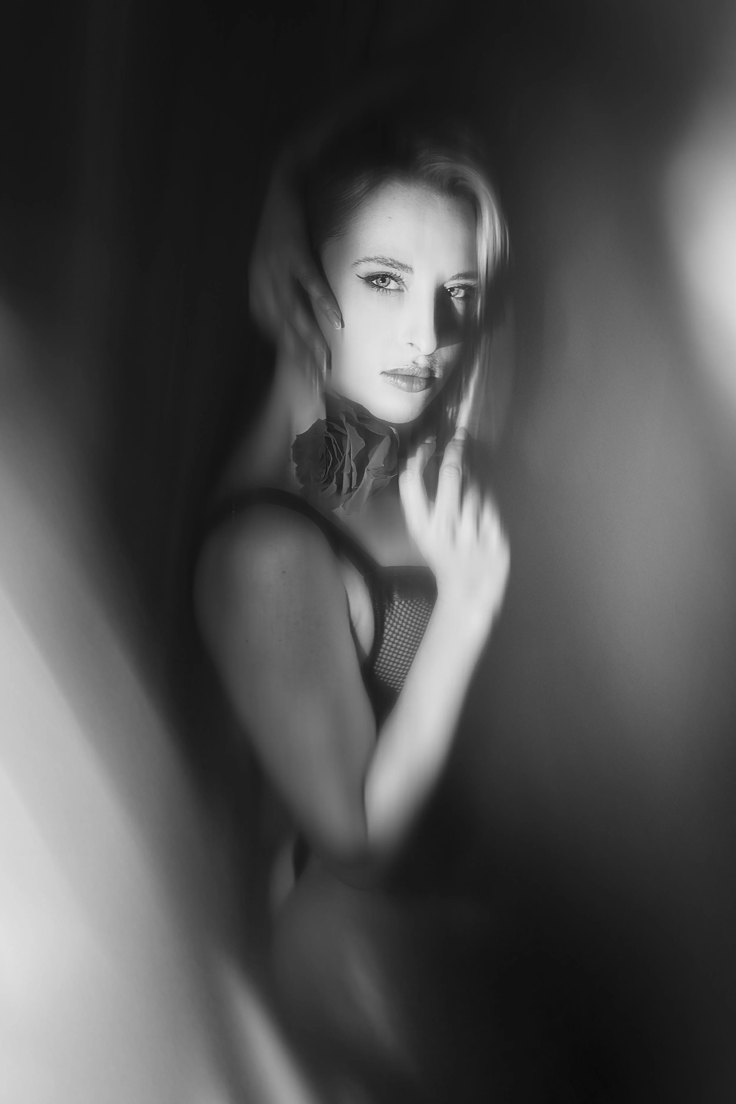 Photo pour séance Portrait Intimiste. Portrait artistique en noir et blanc d'une femme regardant intensément, avec un collier de roses et des vêtements en filet, vue à travers une surface floue.