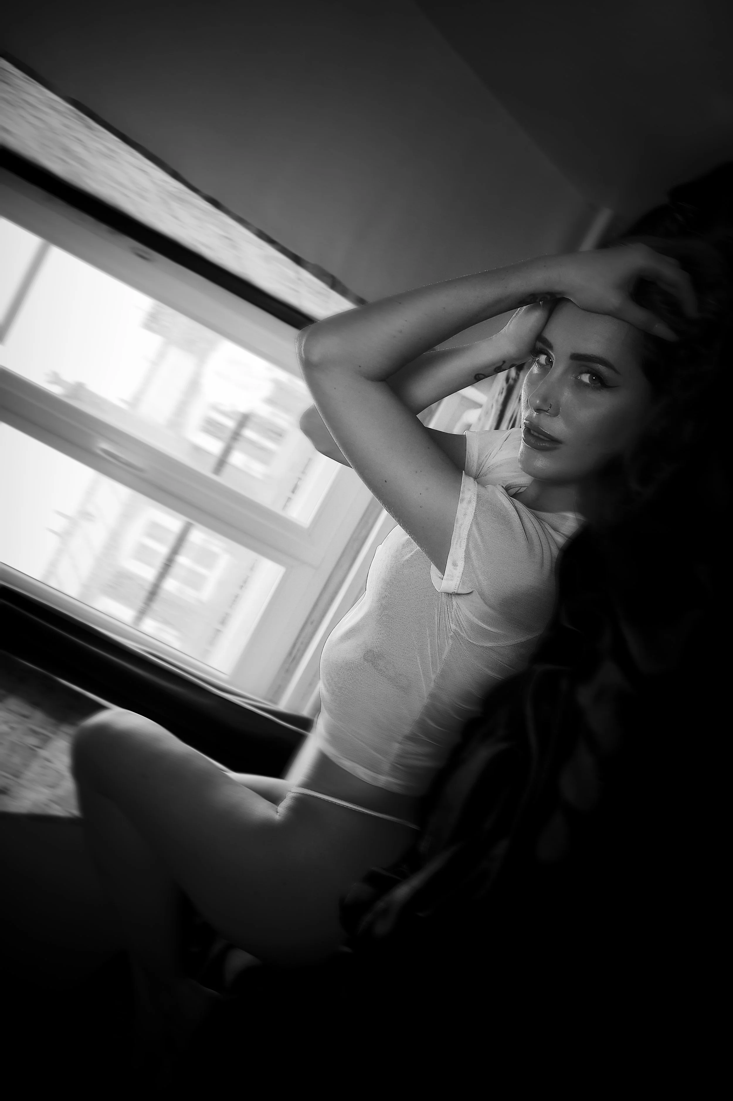 Photo pour séance Portrait Intimiste. Une femme pose devant une fenêtre en noir et blanc, en tenant ses cheveux en arrière. Elle porte un t-shirt blanc moulant et semble regarder l'objectif.