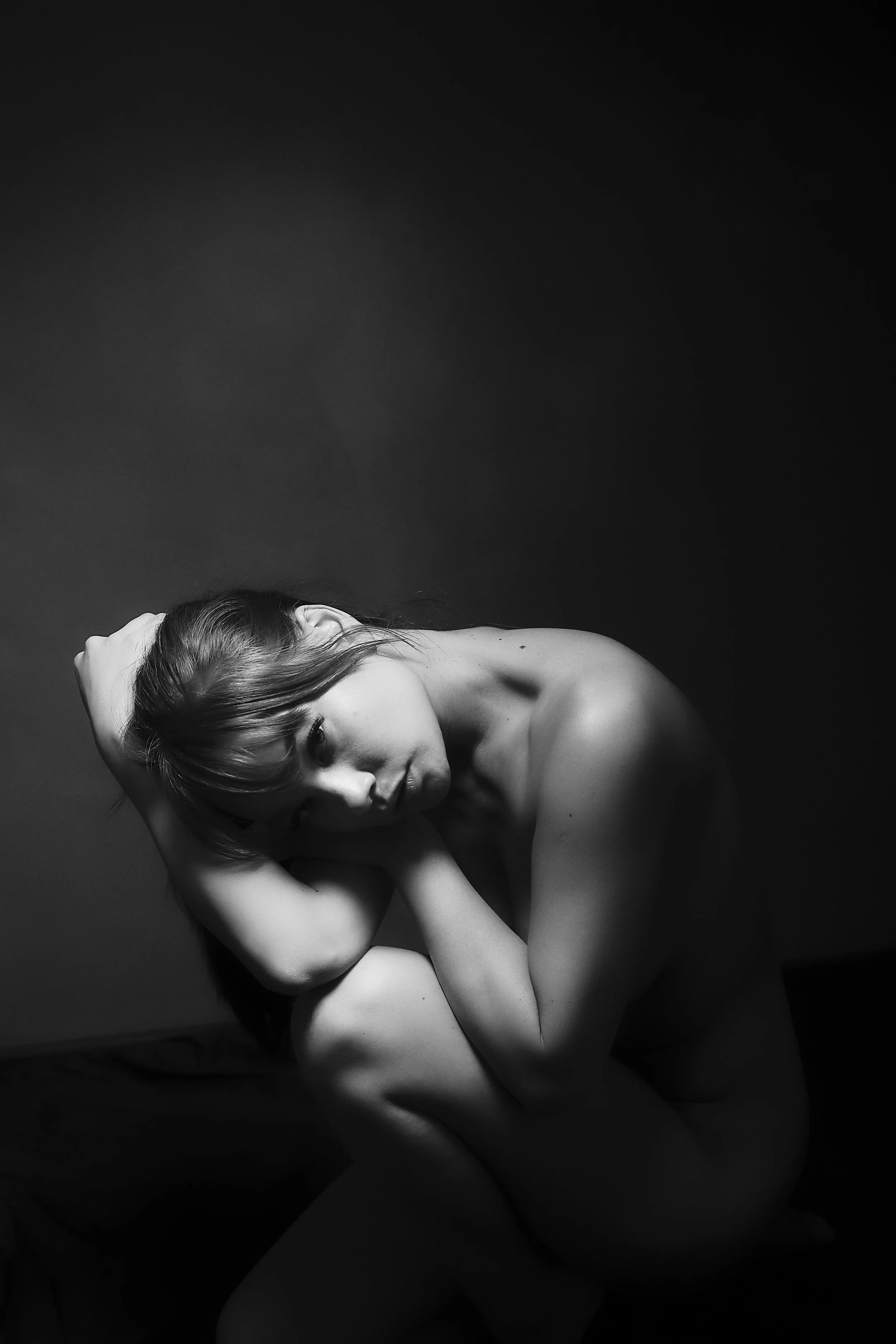 Photo pour séance Portrait Intimiste. Femme nue en position de fœtus, en noir et blanc, avec un éclairage dramatique sur un fond sombre.