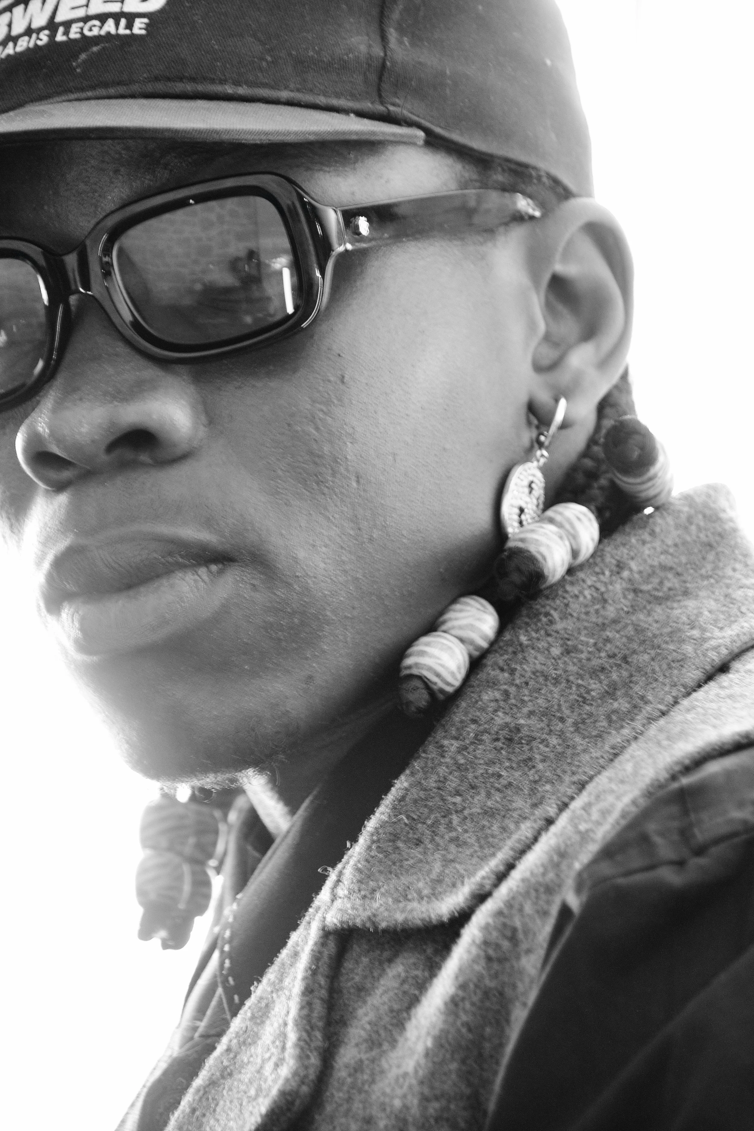 Photo pour séance Portrait Intimiste. Portrait en noir et blanc d'une jeune femme portant des lunettes de soleil, un bonnet et des boucles d'oreilles, avec des perles dans les cheveux, vue de profil.