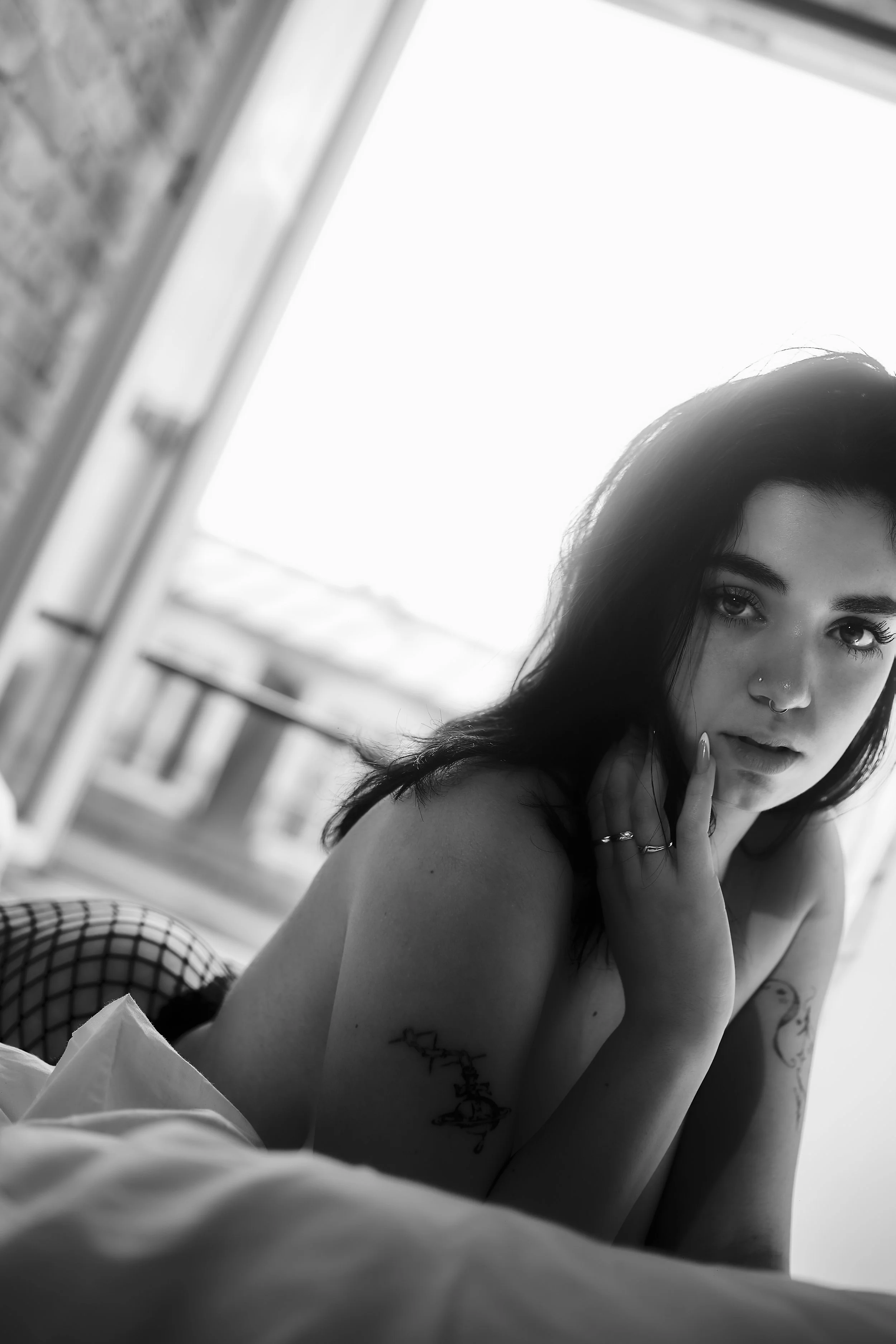 Photo pour séance charme. Jeune femme avec des cheveux noirs, portant un maquillage léger, des tatouages et des bijoux, posant lors d'une séance photo en noir et blanc avec une lumière naturelle provenant d'une fenêtre.