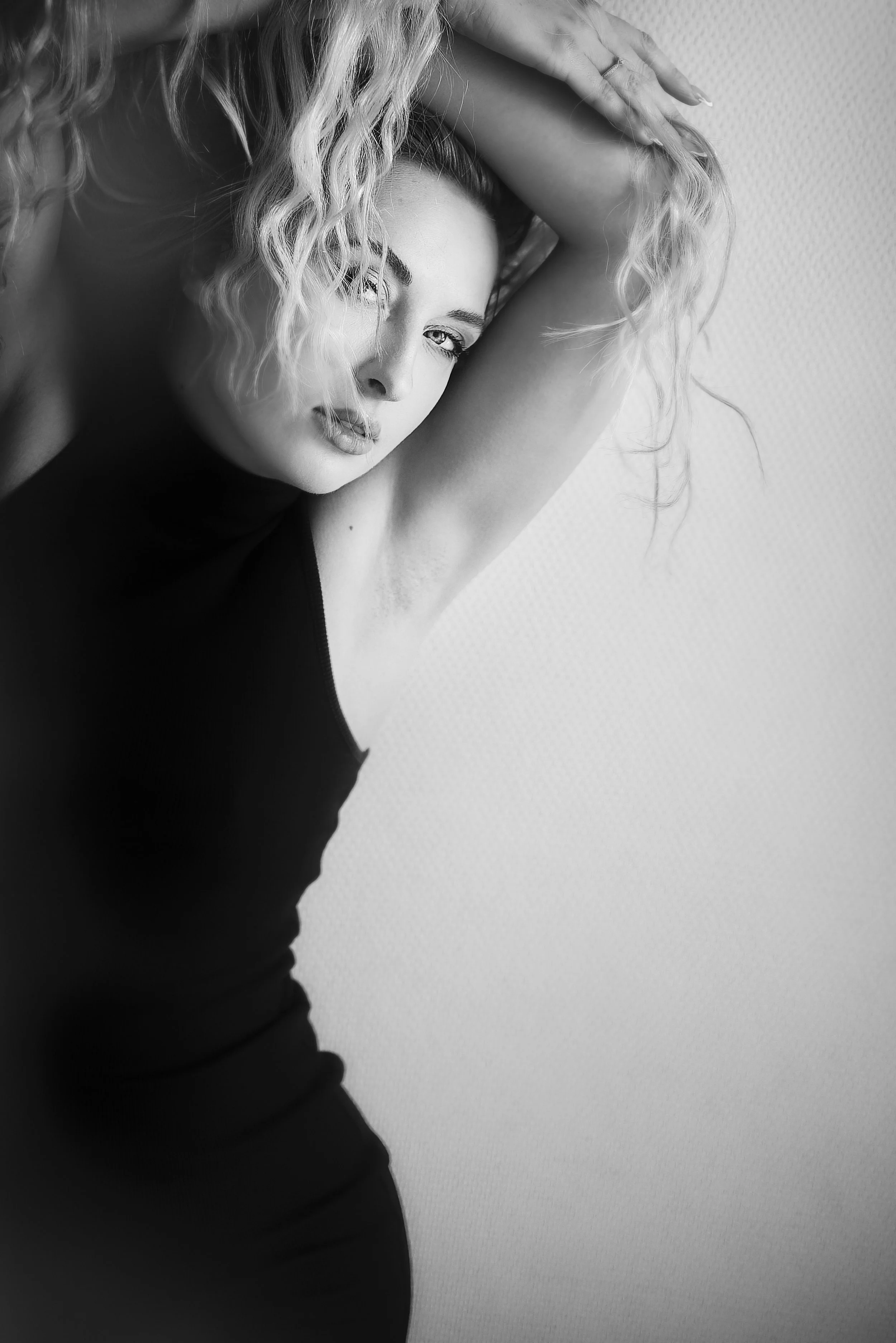 Une femme avec des cheveux bouclés, portant un haut noir, pose devant un fond neutre, regardant vers l'appareil, avec une expression douce et confiante.