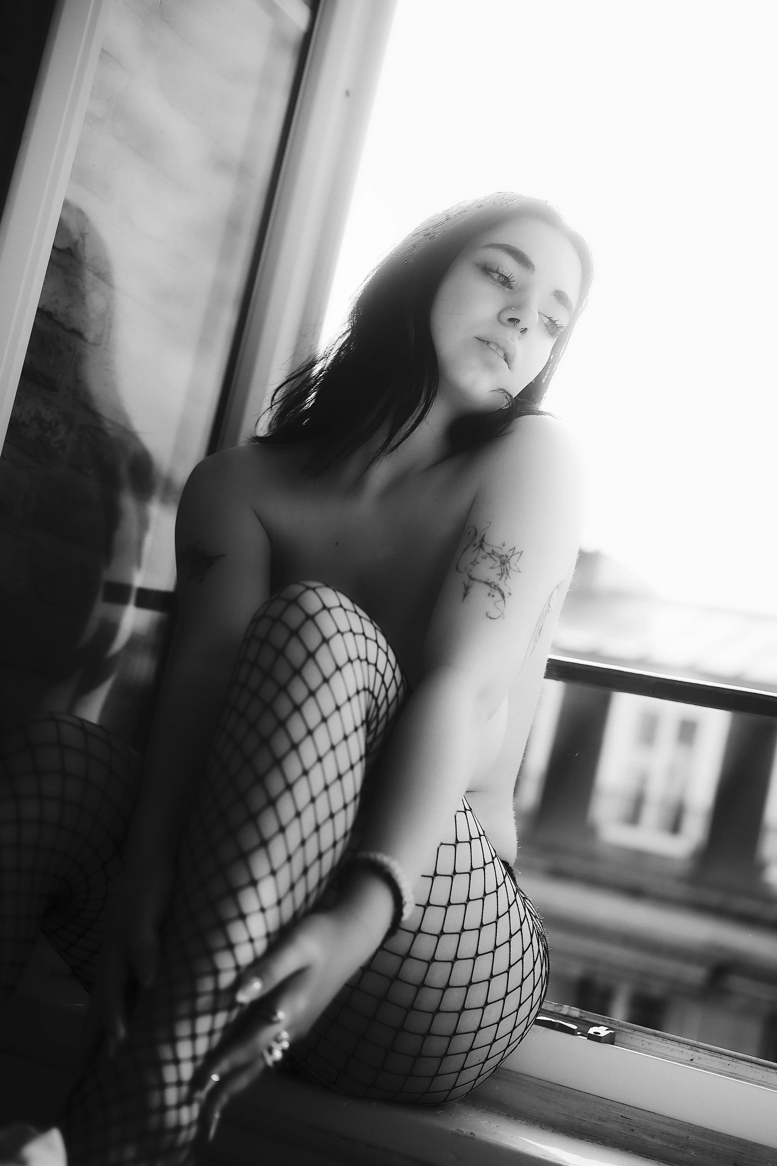 Photo pour séance Boudoir. Une femme aux cheveux noirs, portant des collants en filet, assise près d'une fenêtre, regardant pensivement.